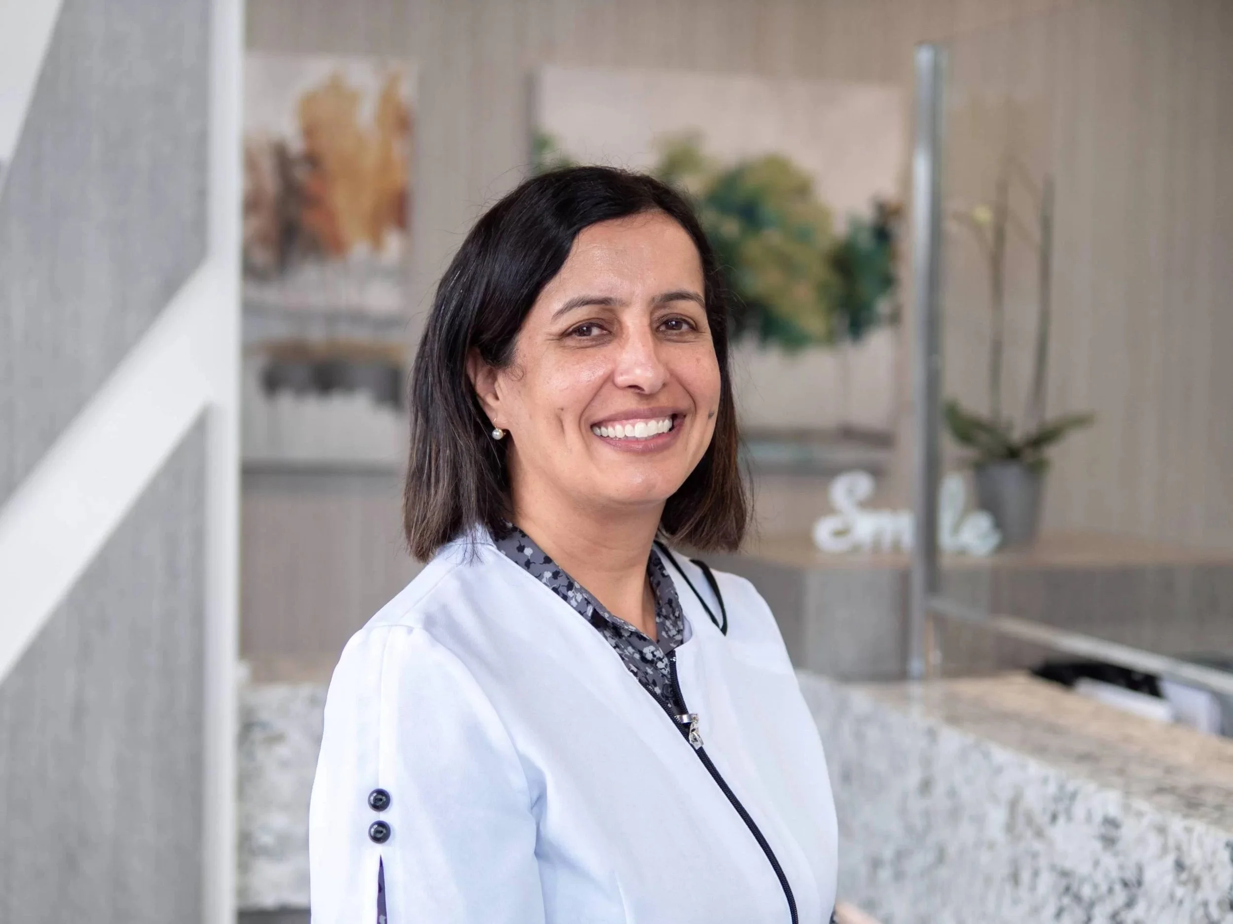 A woman with dark hair, wearing a white medical coat and pearl earrings, smiling indoors with blurred background of artwork and plants.
