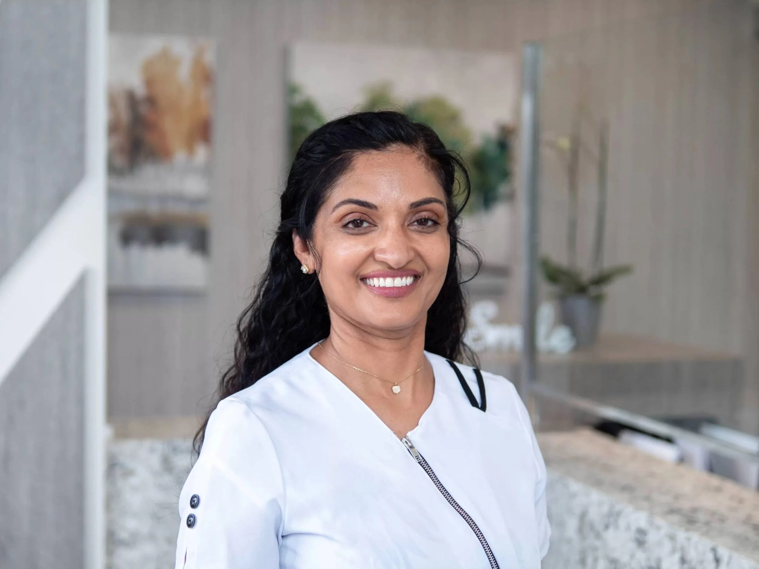 A smiling woman with dark wavy hair, wearing earrings and a white jacket with a zipper, standing outdoors in front of a modern building with glass windows and plants.