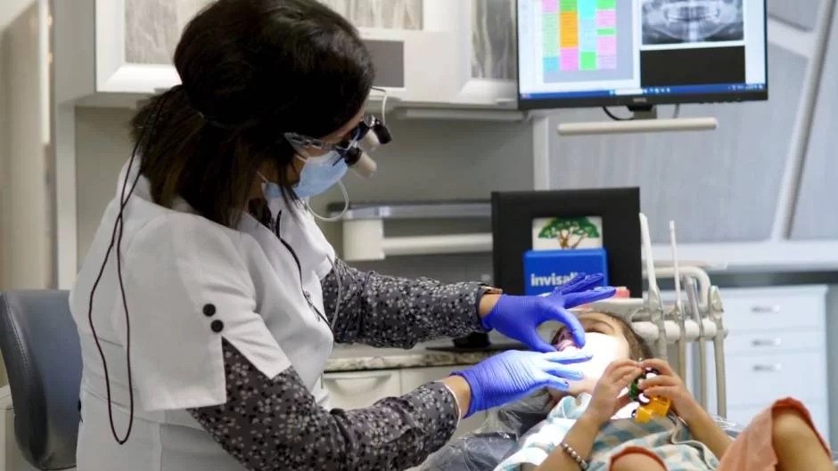 Dentist wearing a mask, gloves, and magnifying glasses examines a child's mouth in a dental clinic.