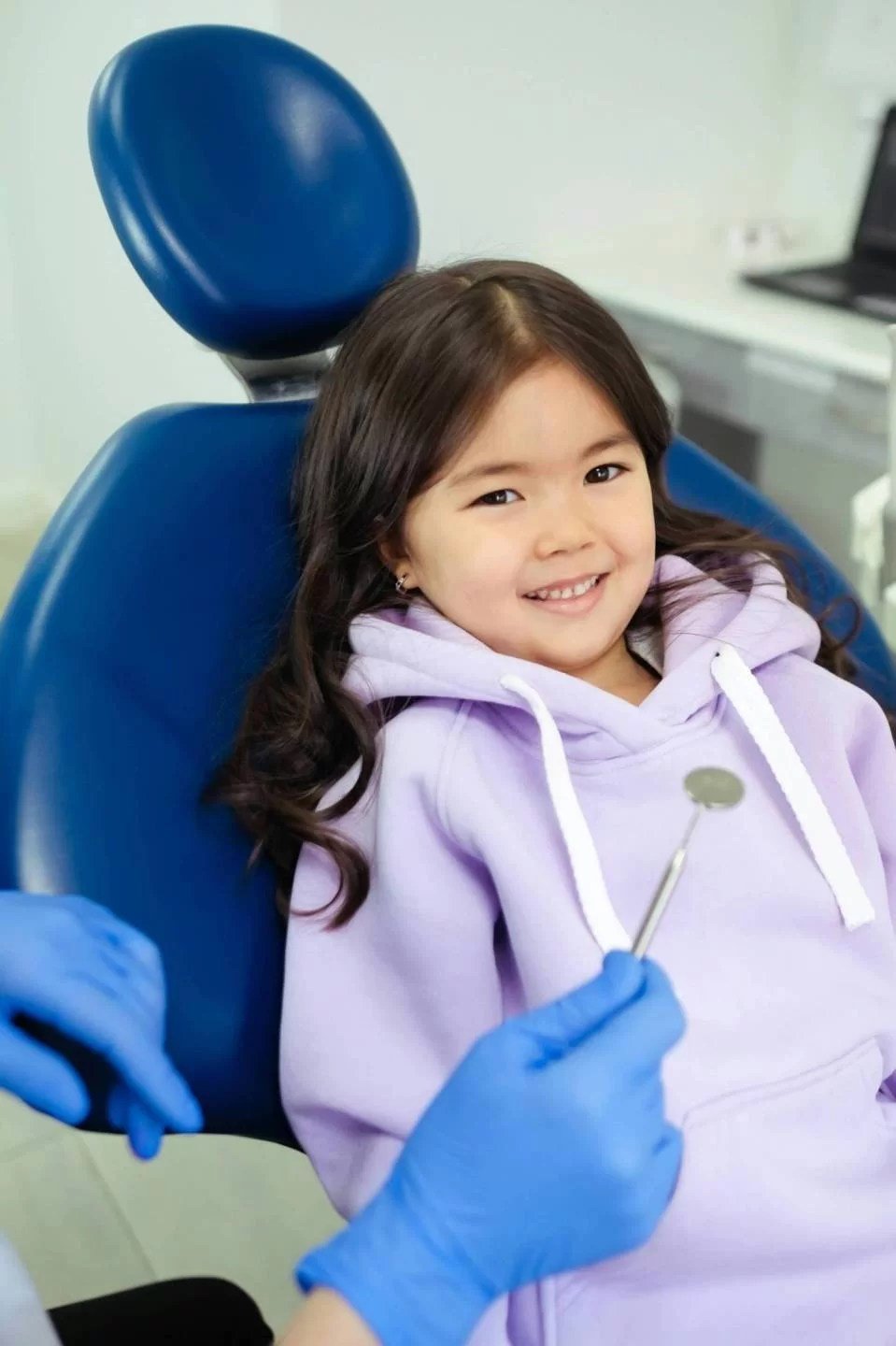 A young girl with long brown hair sitting in a dental chair, smiling at the camera, wearing a lavender hoodie, with a dental mirror held by a person wearing blue gloves.