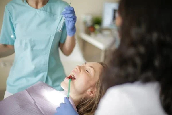 A young girl lying in a dental chair with her mouth open, receiving dental treatment from a dentist wearing a mask and gloves, while another person observes.