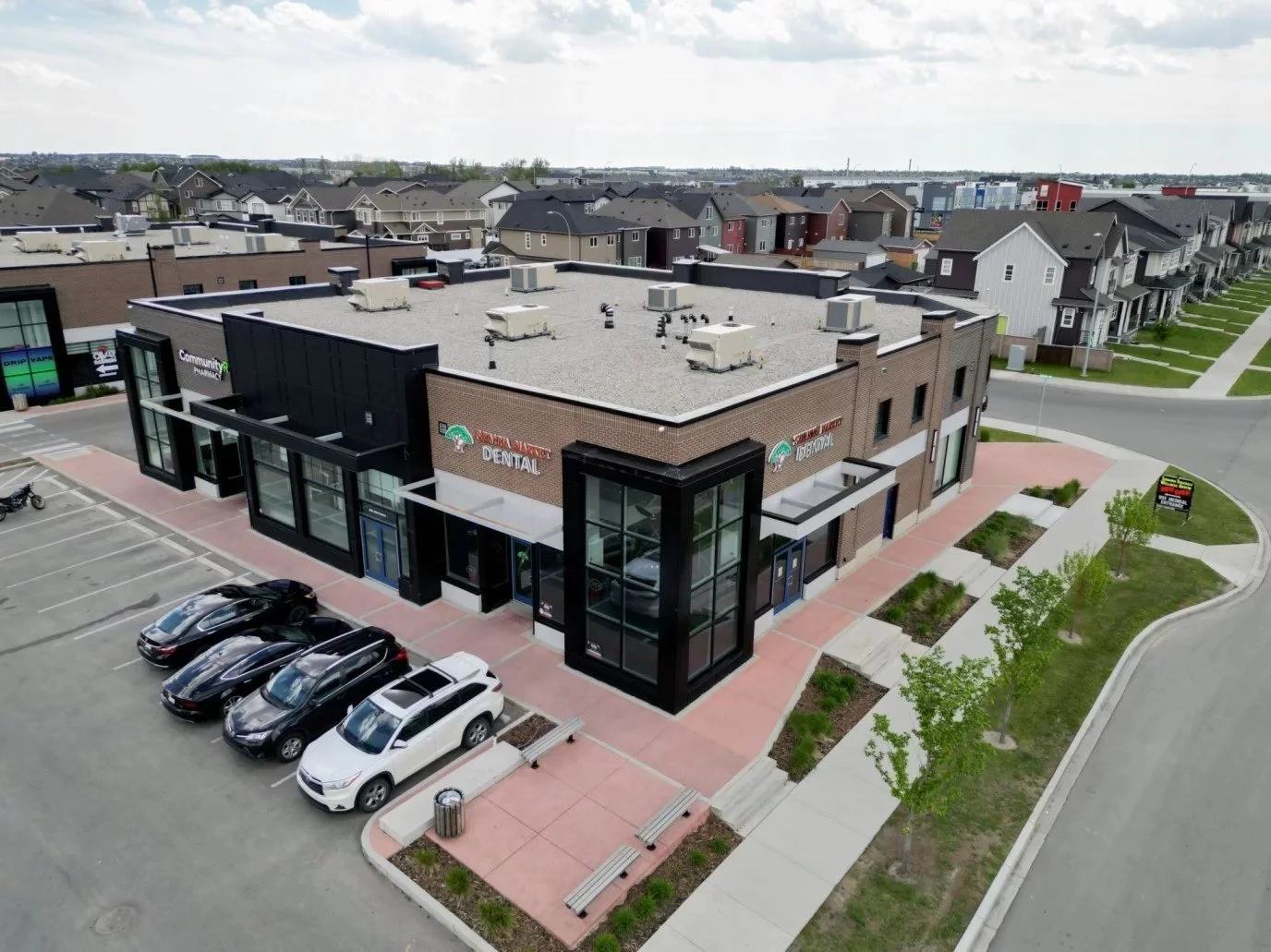 An aerial view of a modern shopping center with a dental office and community pharmacy, surrounded by a parking lot with several cars and a sidewalk with trees and landscaped areas.