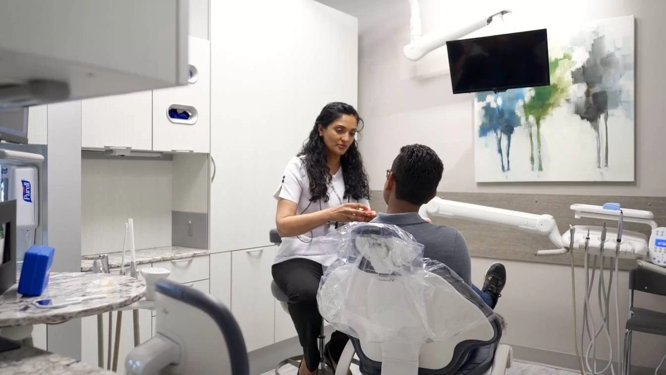 Dentist or dental hygienist attending to a patient in a modern dental clinic. The patient is seated in a dental chair, and the dental professional is standing nearby, holding dental tools. The clinic has white cabinets, a mountain landscape painting, and medical equipment.