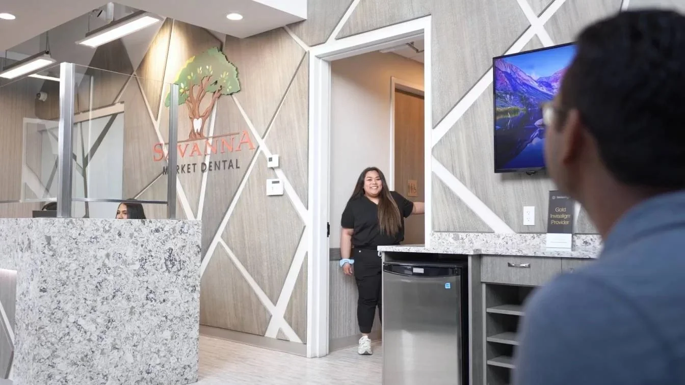 A dental office reception area with a woman smiling and standing in an open doorway, a man sitting in front of a television, and a person behind the reception desk. The wall features a logo with a tree and the words 'Savanna Market Dental.'