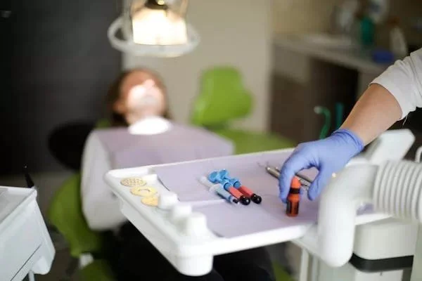 Child lying in a dental chair with a dental professional preparing dental instruments on a tray in a dental clinic.