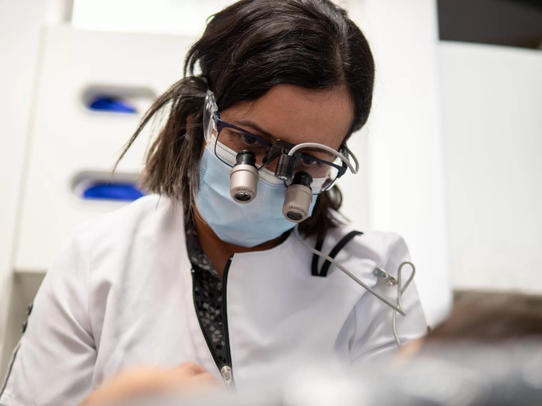 A female scientist wearing magnifying loupes, a face mask, and a white lab coat, working in a laboratory with cabinets and equipment in the background.
