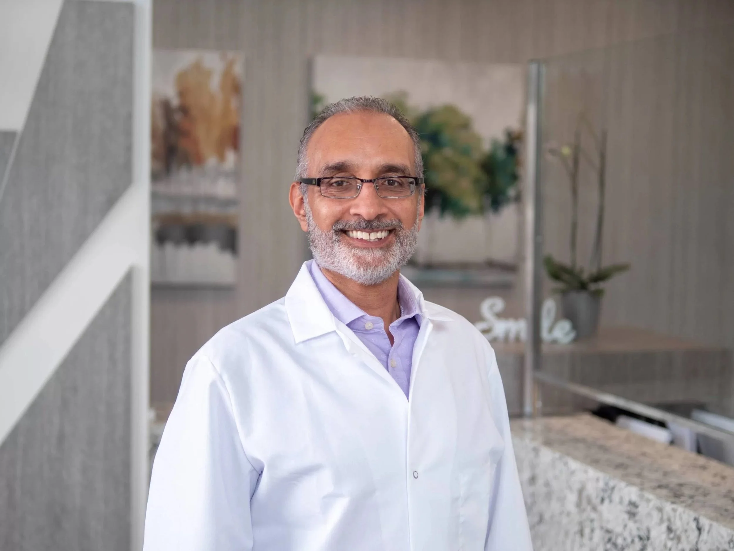 A middle-aged man with glasses and a beard, smiling, wearing a white lab coat and a lavender shirt, standing outdoors with modern building and plants in the background.