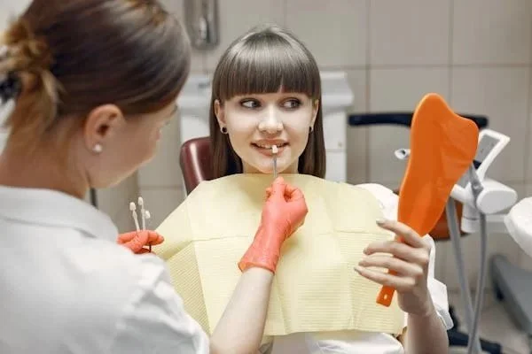 A young girl at a dental appointment holding a mirror and looking at her teeth, while a dentist or dental hygienist examines her mouth.