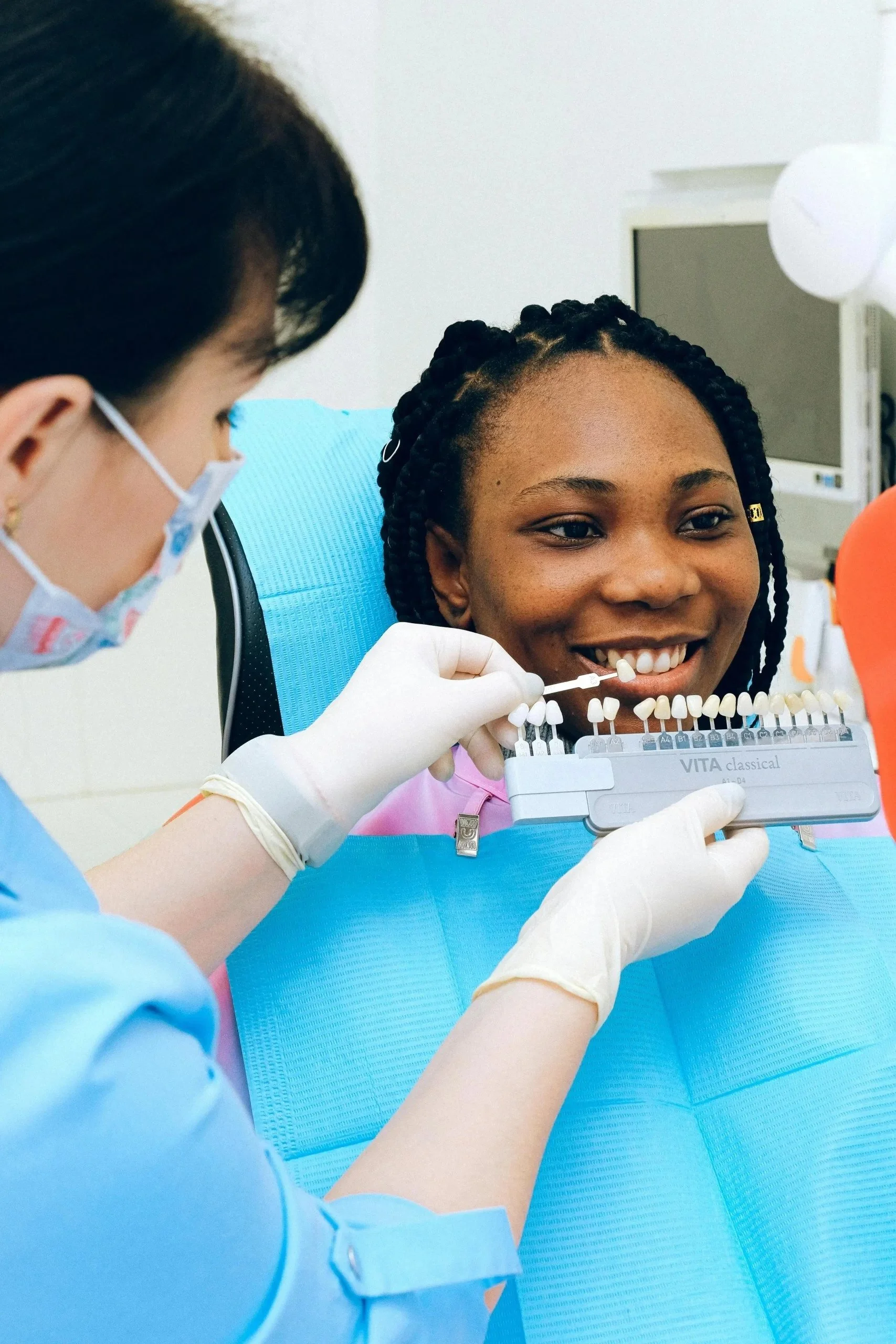 A woman with braids smiling at the dentist during a dental check-up, with dental shade guides in her mouth.