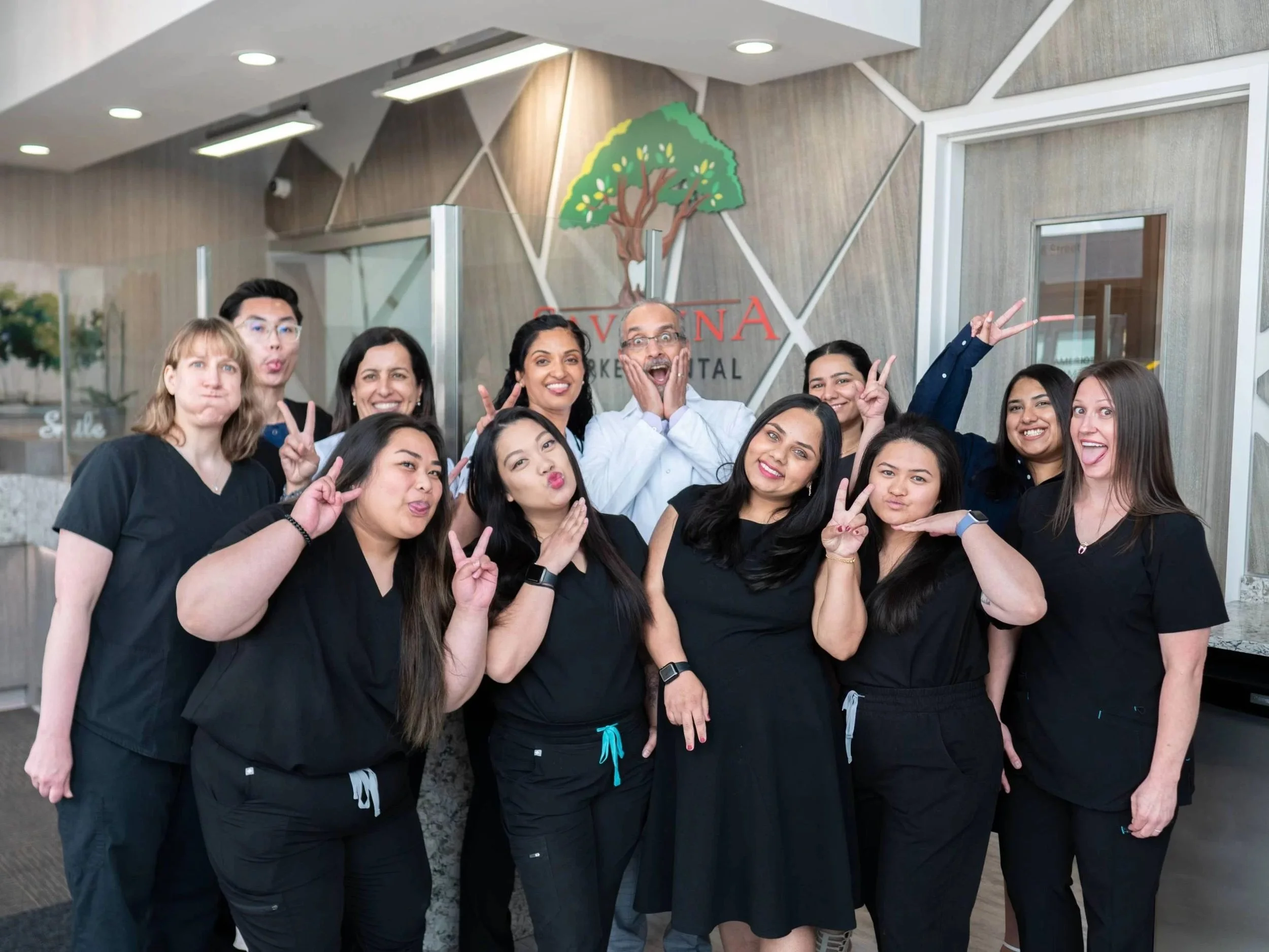 A group of diverse people in scrubs posing and smiling in an indoor setting with a wooden wall and a tree logo behind them.