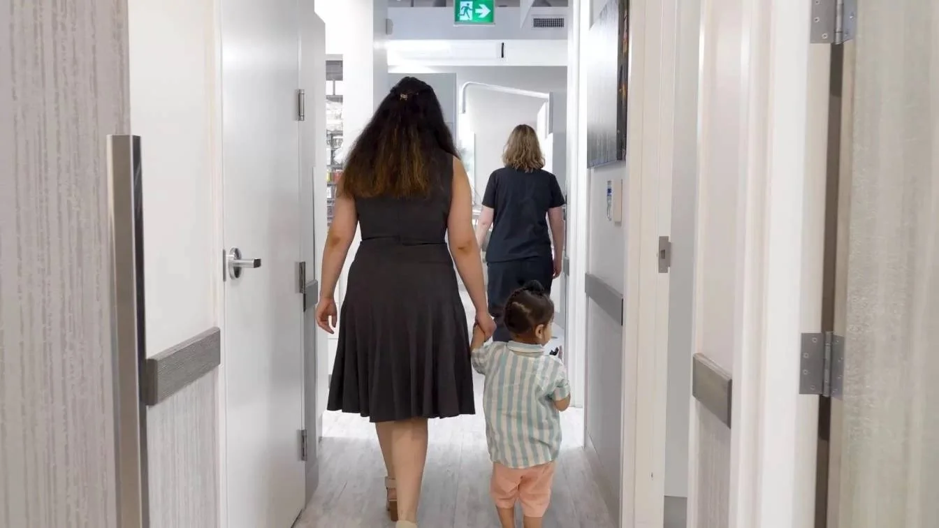 A woman holding a child's hand walking down a hospital corridor with healthcare workers visible in the background.