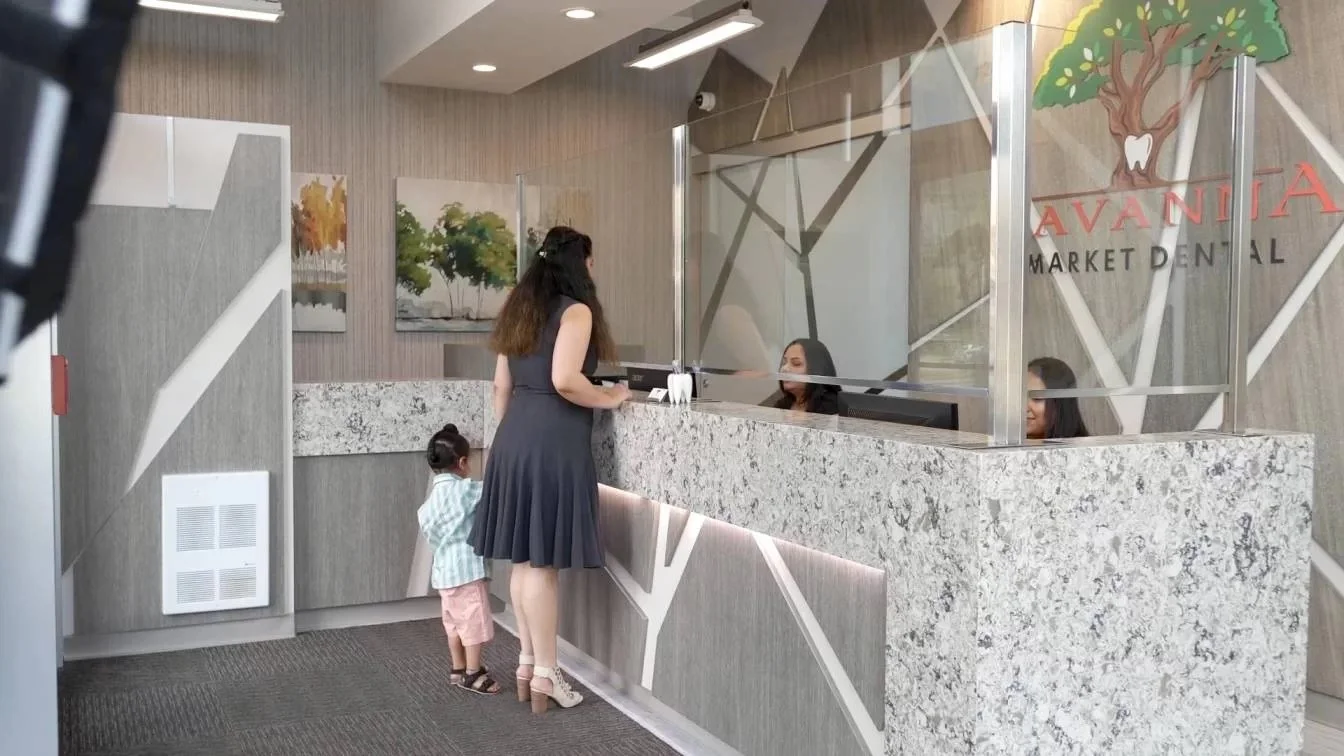 A woman with a toddler at a dental clinic reception desk. The woman is standing and talking to a receptionist behind a glass partition, while the child looks on. The clinic's name, Avantia, and the words 'Market Dental' are visible on the glass. Artworks of trees and a landscape are hung on the wall behind the reception area.