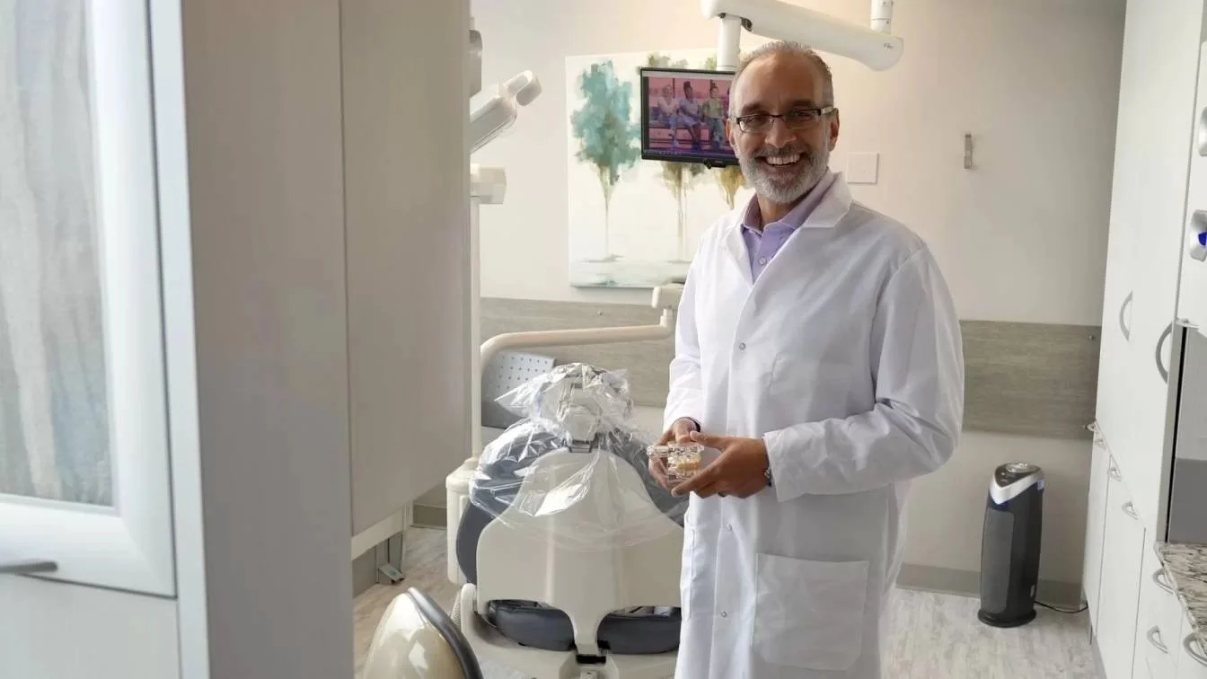 A dentist in a white coat standing in a dental office holding a dental mold, smiling at the camera.