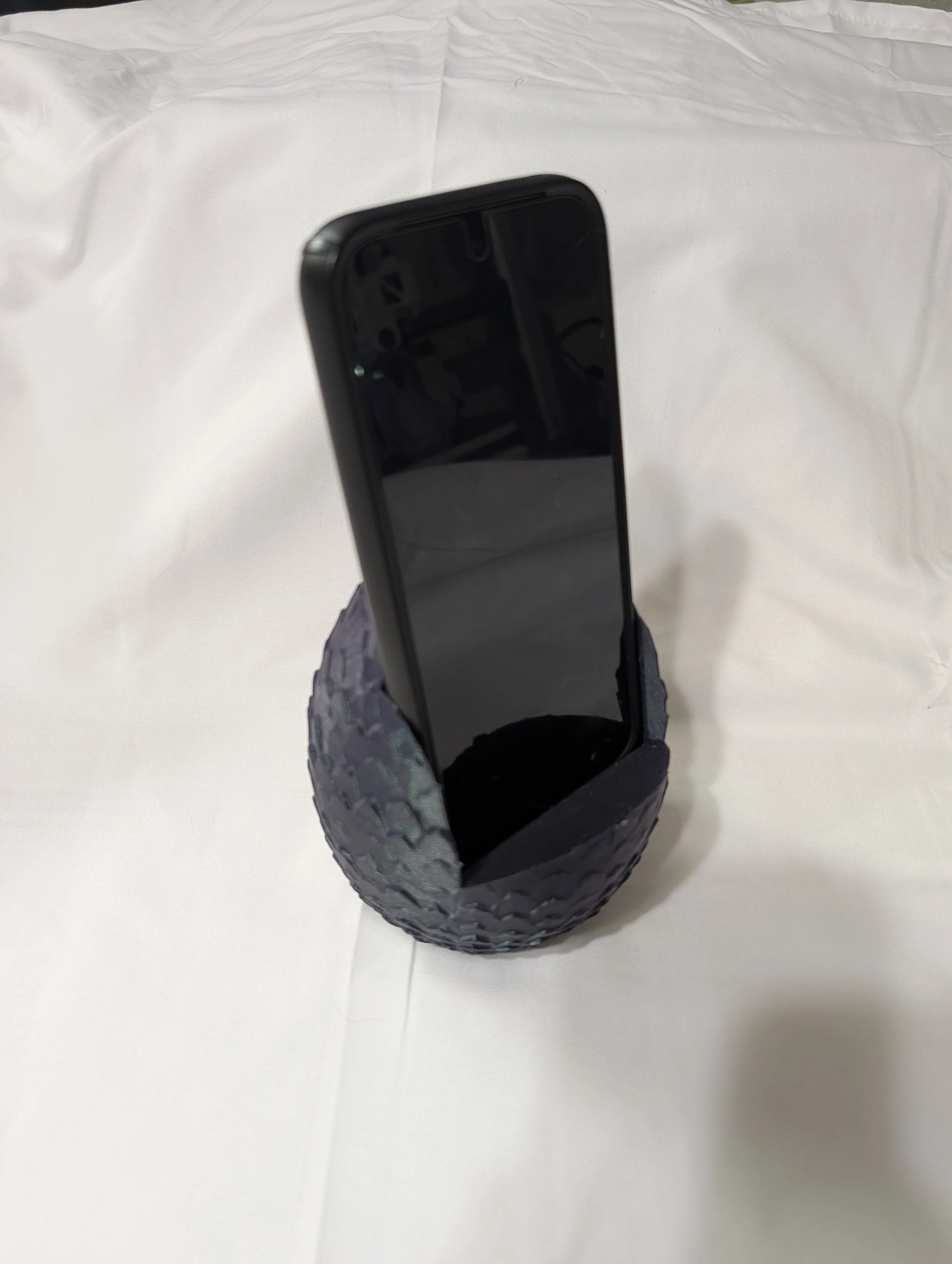 A smartphone with a black screen propped up inside a textured black container on a white surface with crumpled white paper in the background.