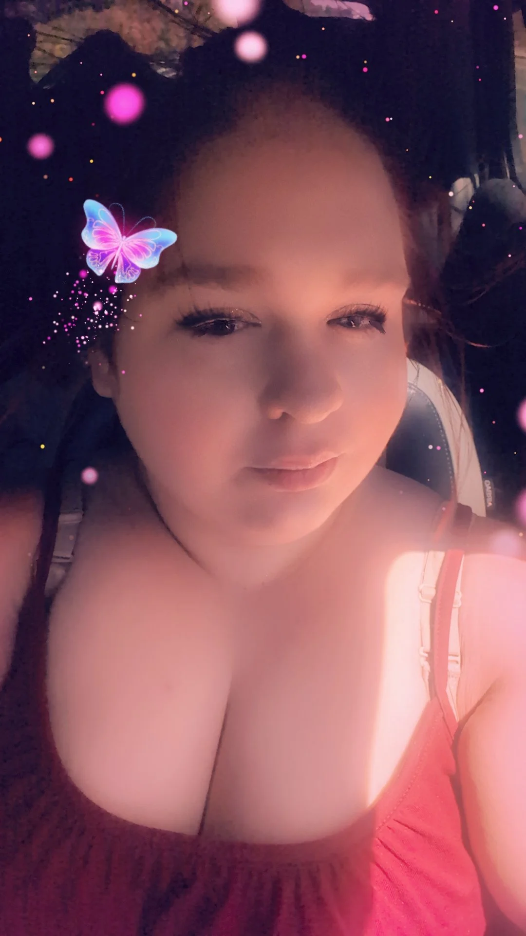 A close-up selfie of a woman with light skin, closed eyes, and makeup, lying down with her arm resting on her forehead, wearing a red top, viewed from above, with a butterfly snapchat filter and pink sparkles overlay.
