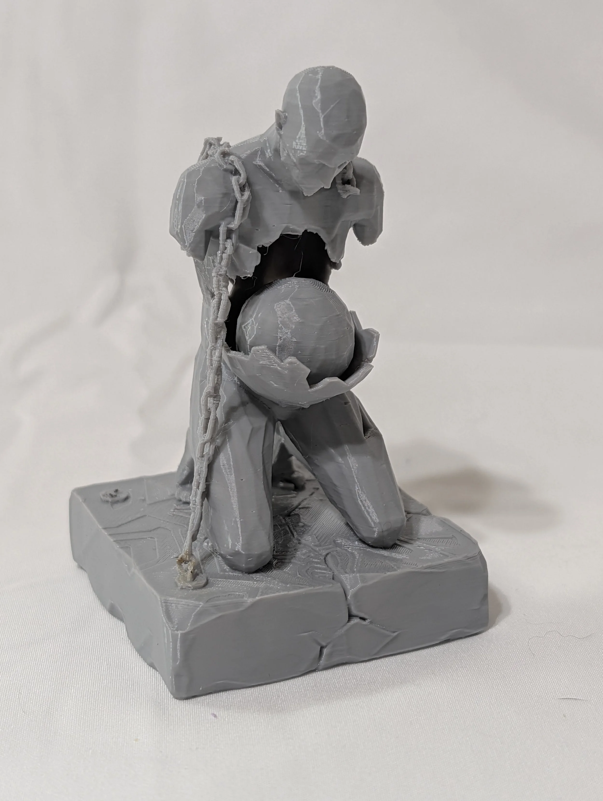 A gray 3D-printed sculpture of a person kneeling on a platform, holding a ball, with chains around their shoulders, against a plain white background.