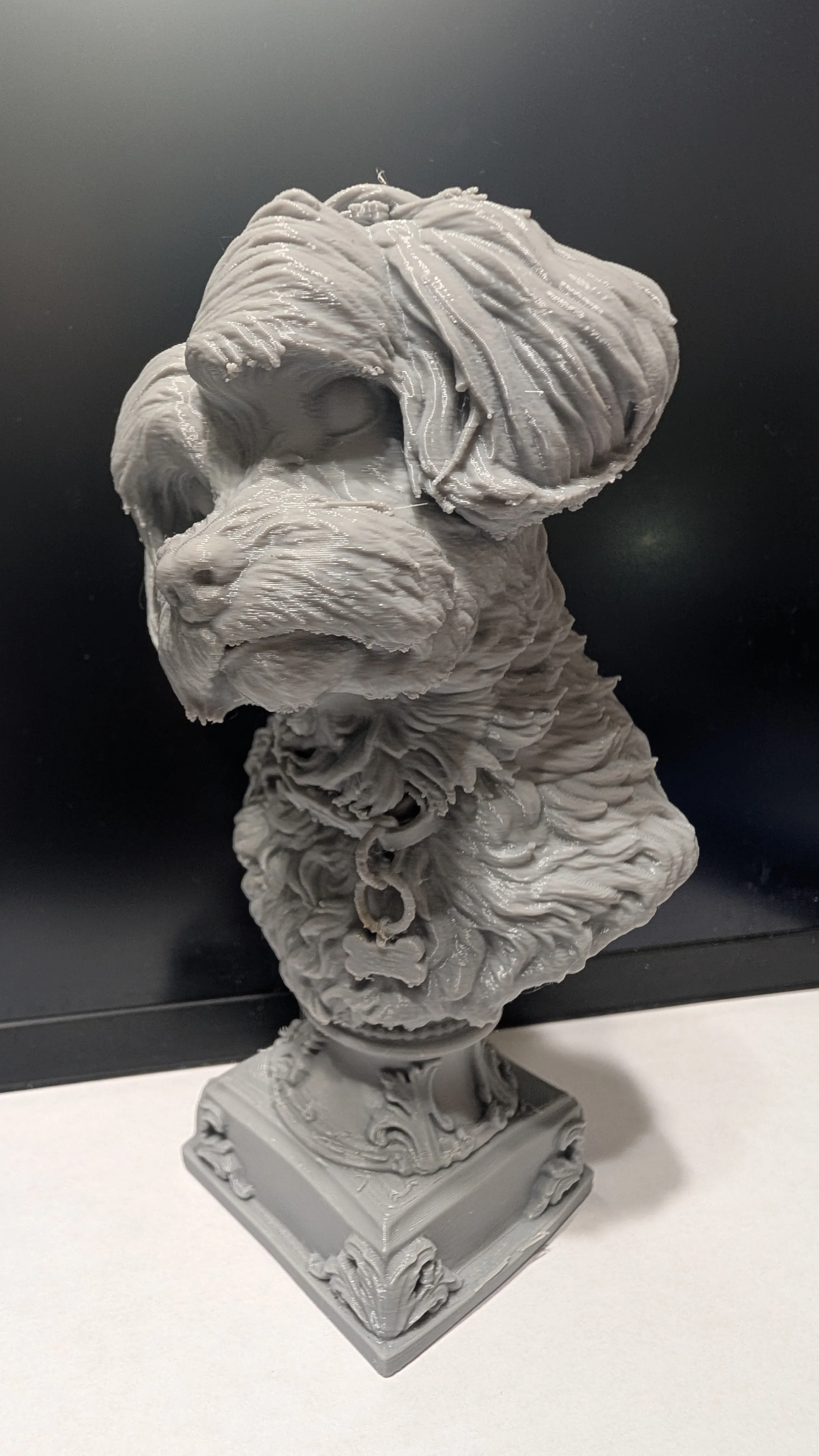 A gray bust sculpture of a dog with fur detailed texture, mounted on a decorated square pedestal.