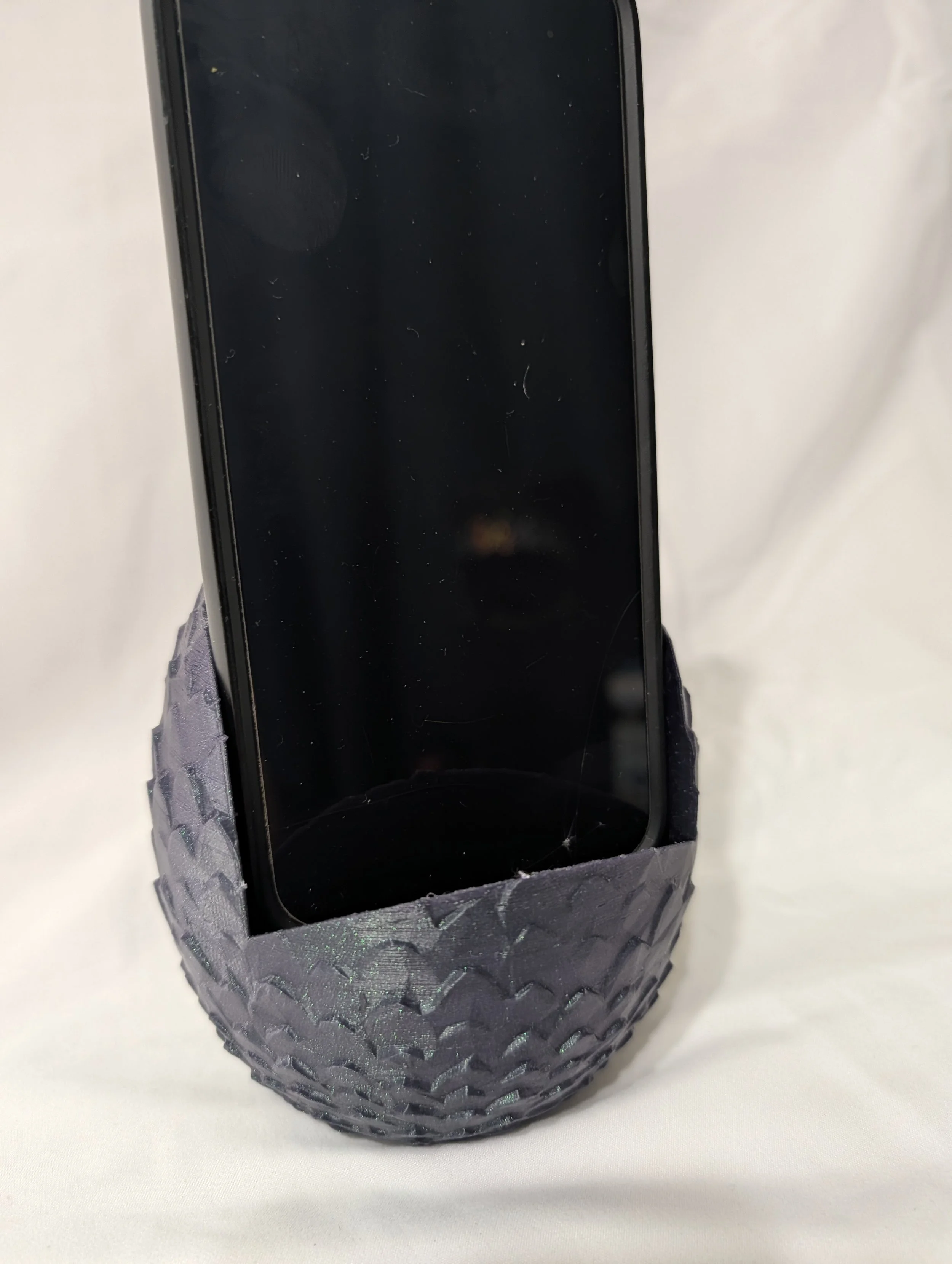 A smartphone with a black screen in a silver case, sitting on a paper towel roll wrapped in silver metallic tape, on a white surface.