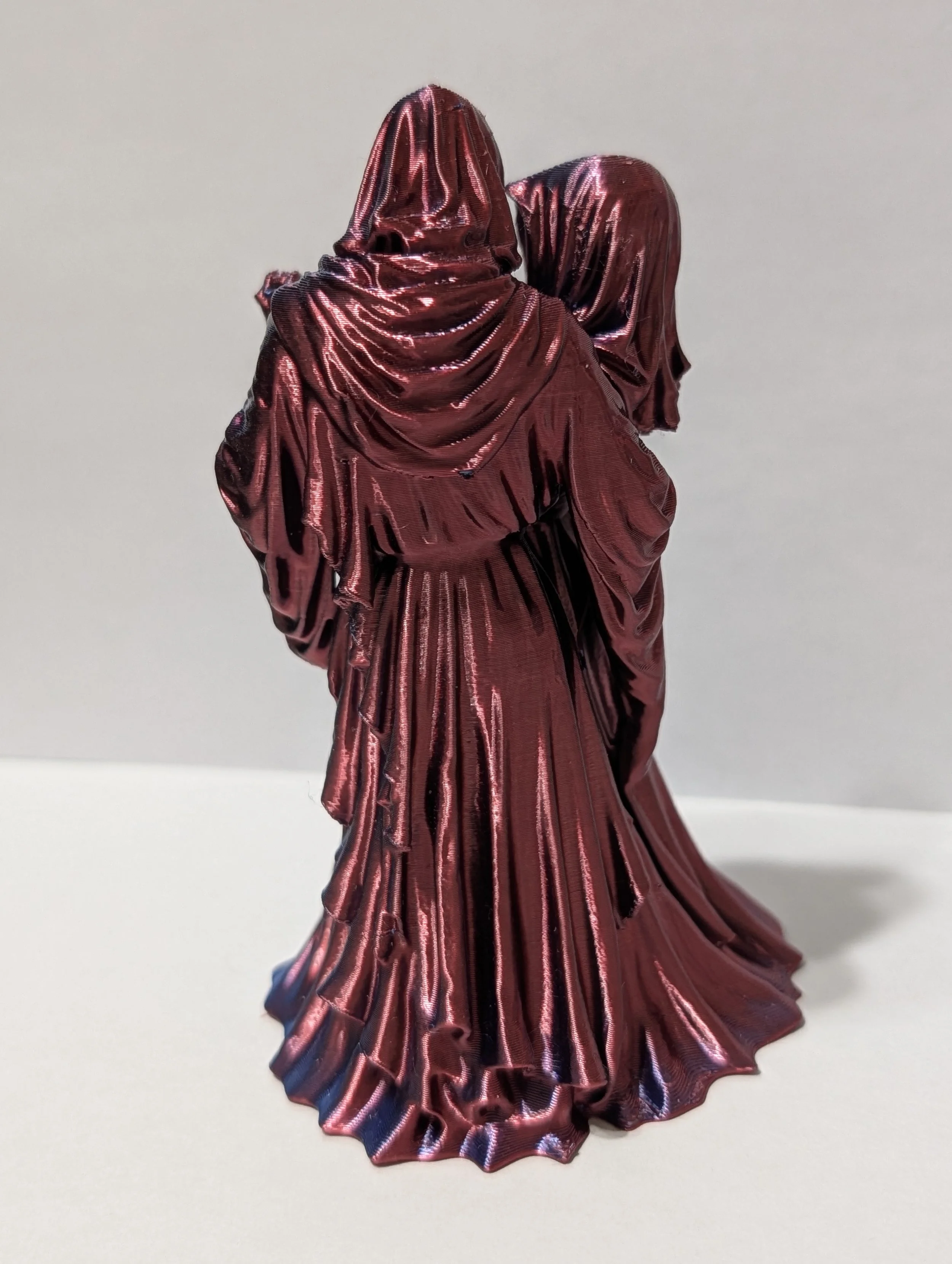 A decorative figure-shaped object resembling a woman in a long flowing dress and hood, made of shiny, metallic, reddish-pink material with pleats and folds.