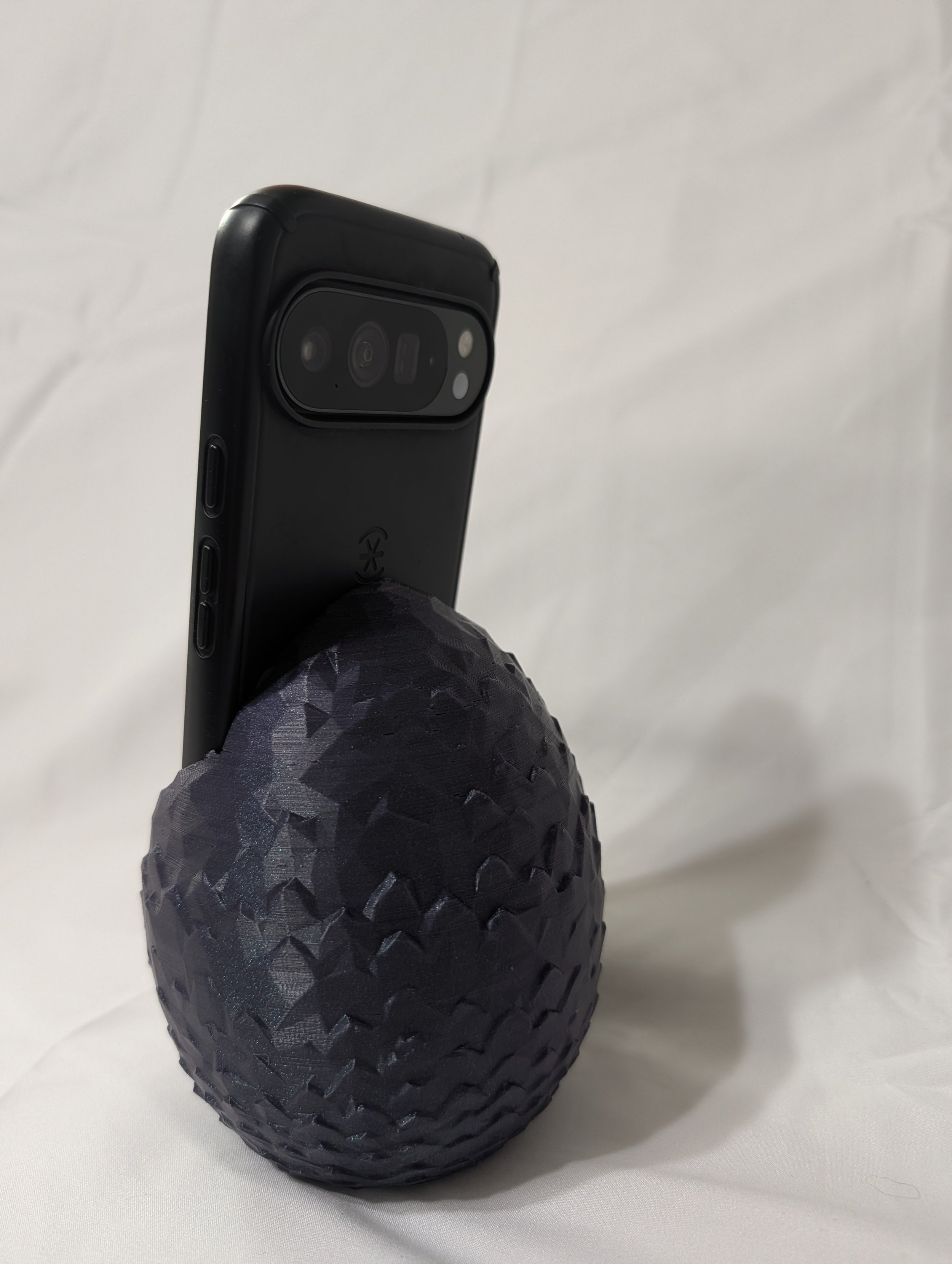 A black rugged smartphone in a protective case with a textured geometric pattern, resting on a white surface.