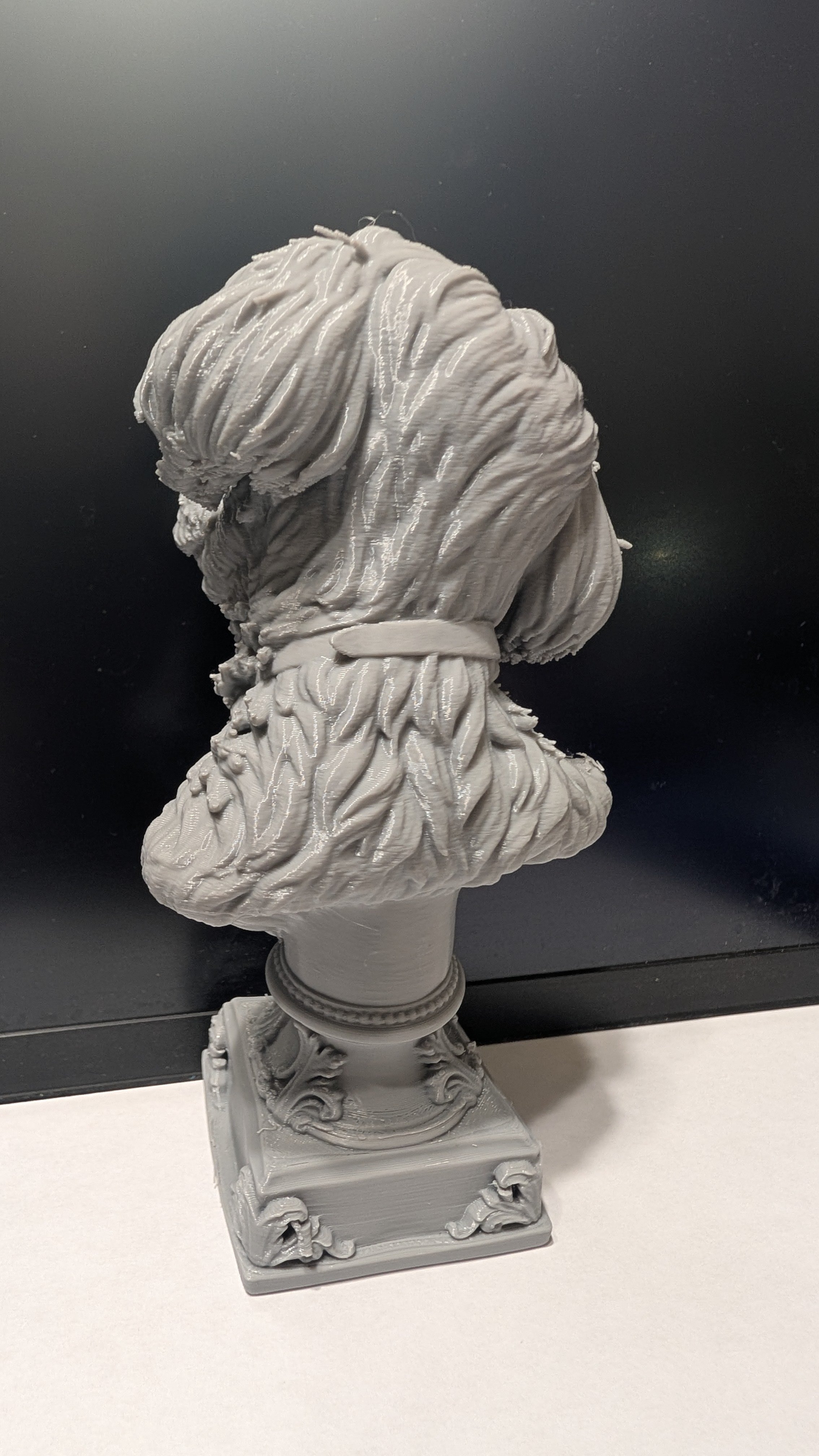 Gray sculpture bust of a woman with wavy hair, mounted on a decorative base.
