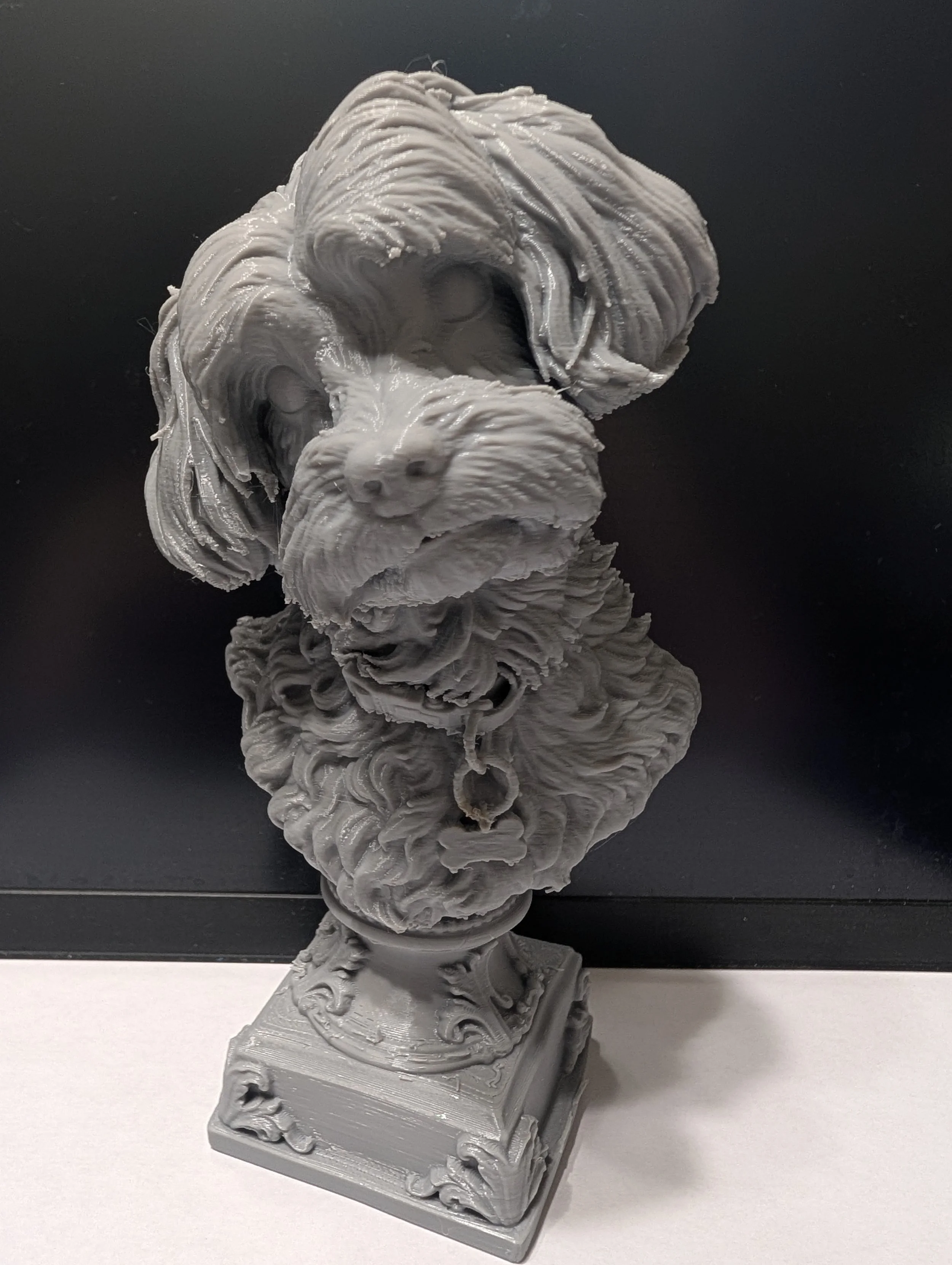 A 3D-printed sculpture of a dog, resembling a retriever, made of gray plastic, placed on a rectangular base with intricate designs, with a black background in one image and a white background in the other.