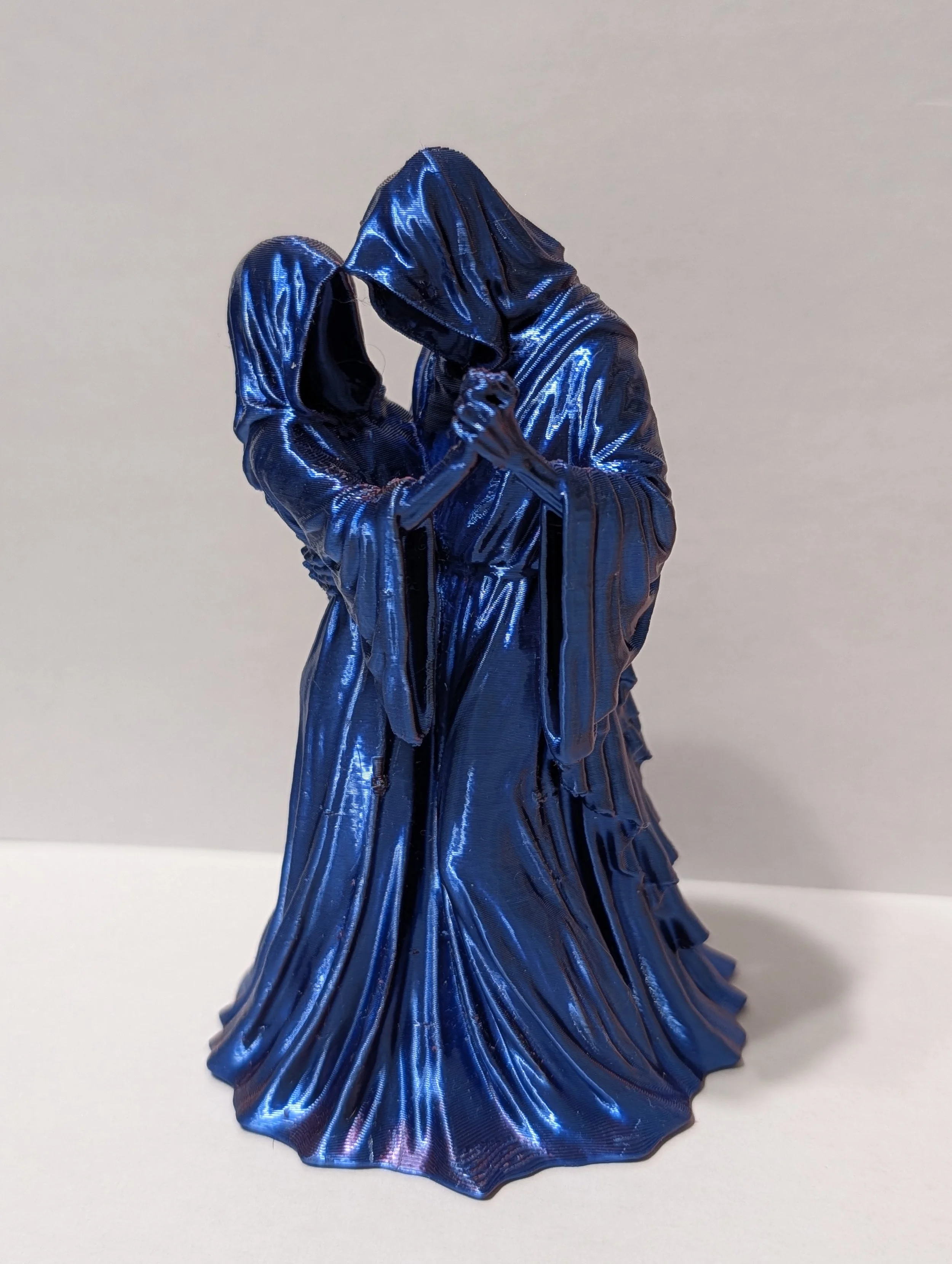 A blue metallic dress sculpture with a flowing, pleated design and a hood, displayed on a white surface against a plain background.