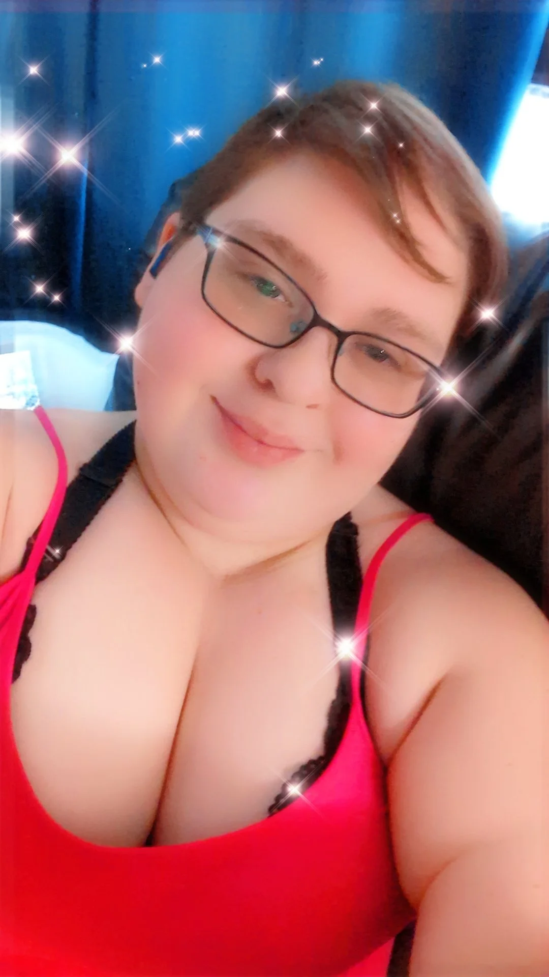 Close-up of a smiling woman with glasses, lying on a bed with a blue headboard, wearing a red tank top, with sparkle effects added to the image.