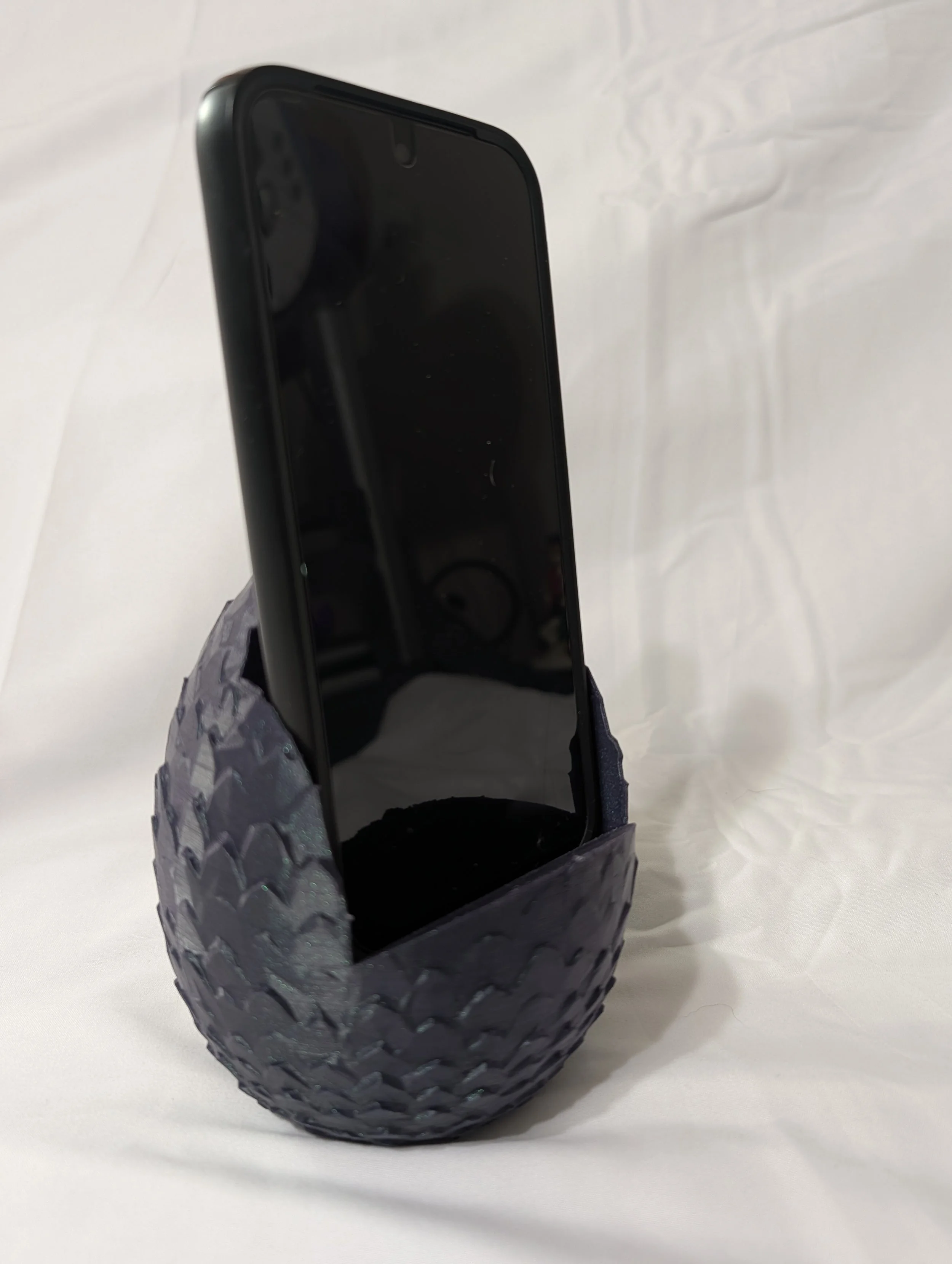 A smartphone placed upright, held in a rubber tire-like holder with a textured pattern. The phone's screen is black and dusty.