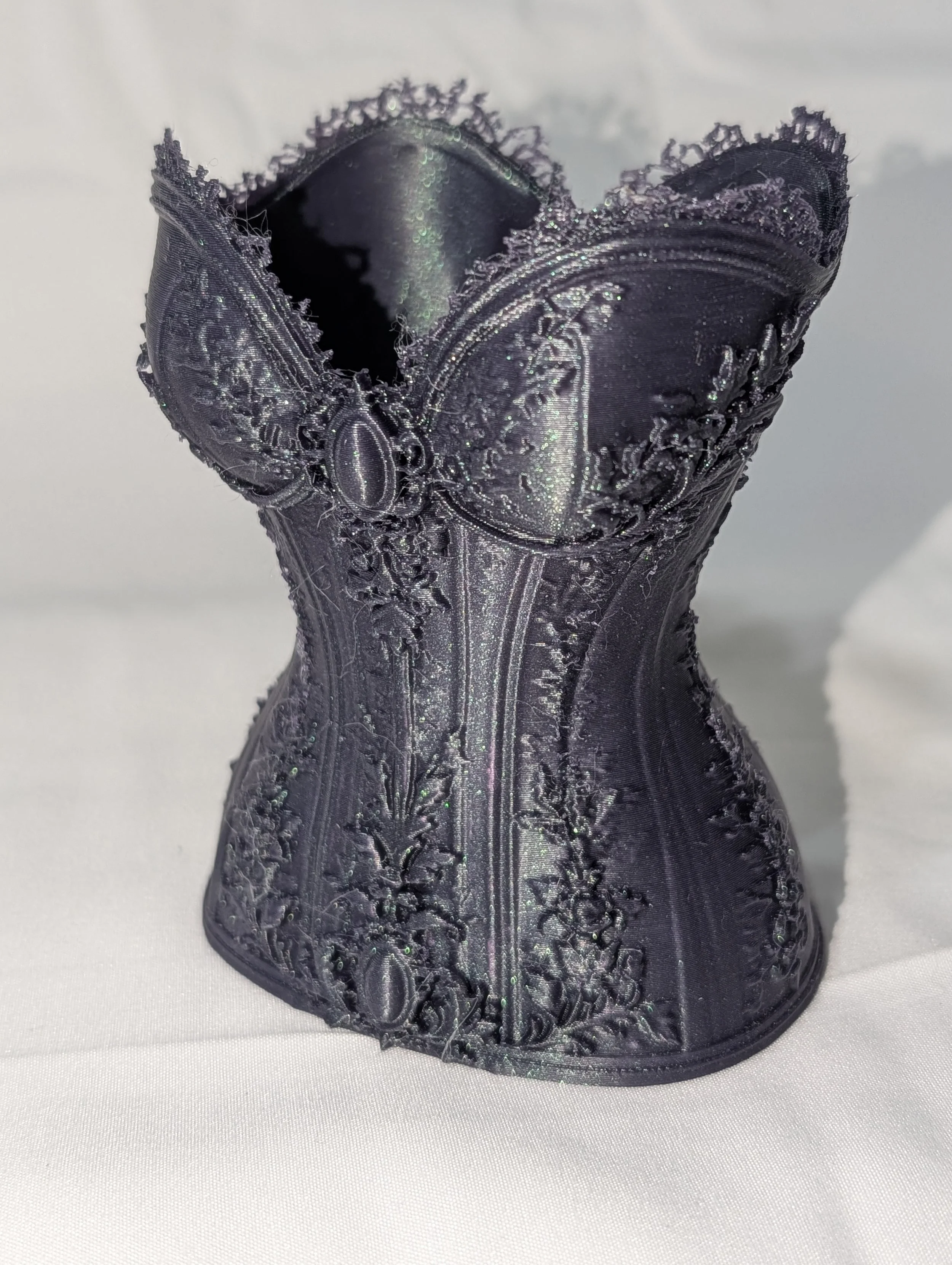 A heart-shaped sculpture made from torn, dark-colored, ornate fabric with glitter and embroidery details, placed on a white surface.