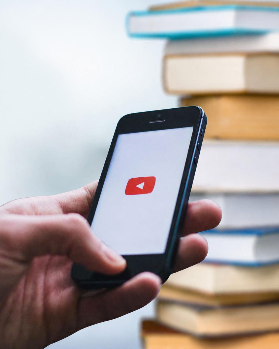 Person holding a smartphone displaying the YouTube logo on the screen, with a background of stacked books.