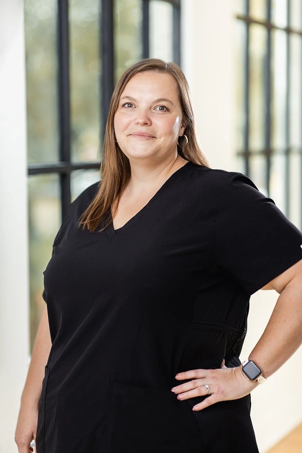 A woman with light skin, long brown hair, and hoop earrings, standing indoors in front of large windows, wearing a black short-sleeved shirt, a watch, and a ring, looking at the camera with a slight smile.