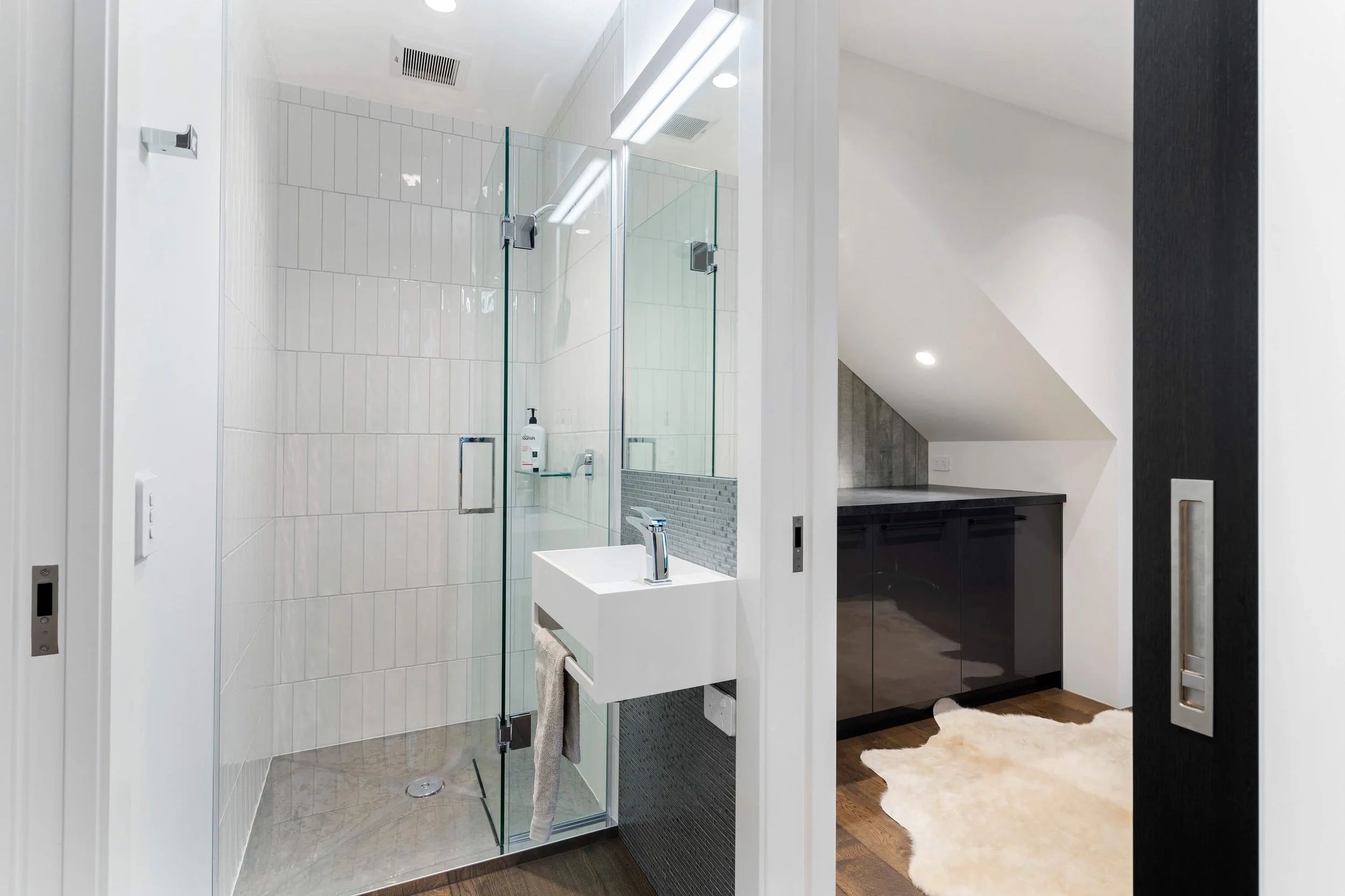 Modern bathroom with glass shower enclosure, white tiles, a small sink, and a dark vanity cabinet with a gold handles. The bathroom has a sloped ceiling and a fluffy white rug on a wooden floor.