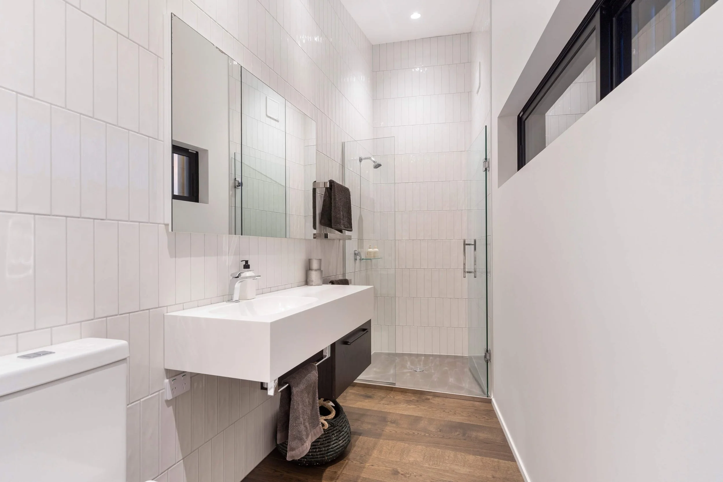 Modern bathroom with white tiled walls, a wall-mounted sink, a large mirror, a glass shower enclosure with a rain shower head, dark towel, small window, and wooden flooring.