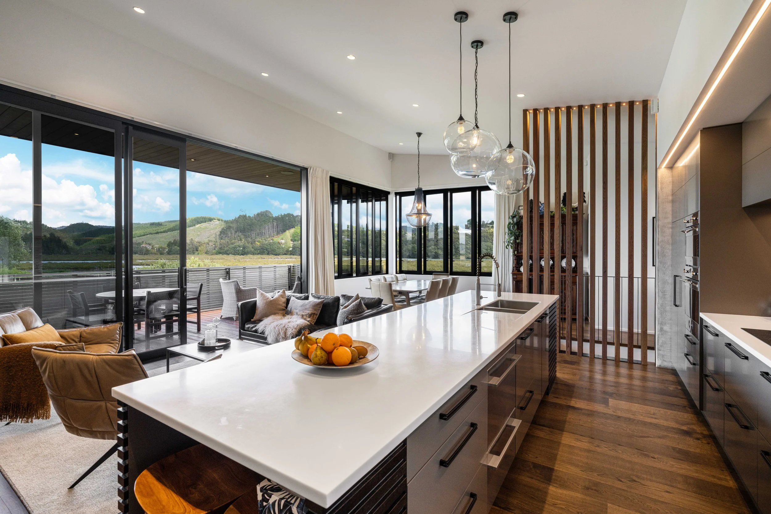Modern open-concept kitchen and living area with large windows, hanging light fixtures, a white marble kitchen island, and a view of a scenic outdoor landscape.