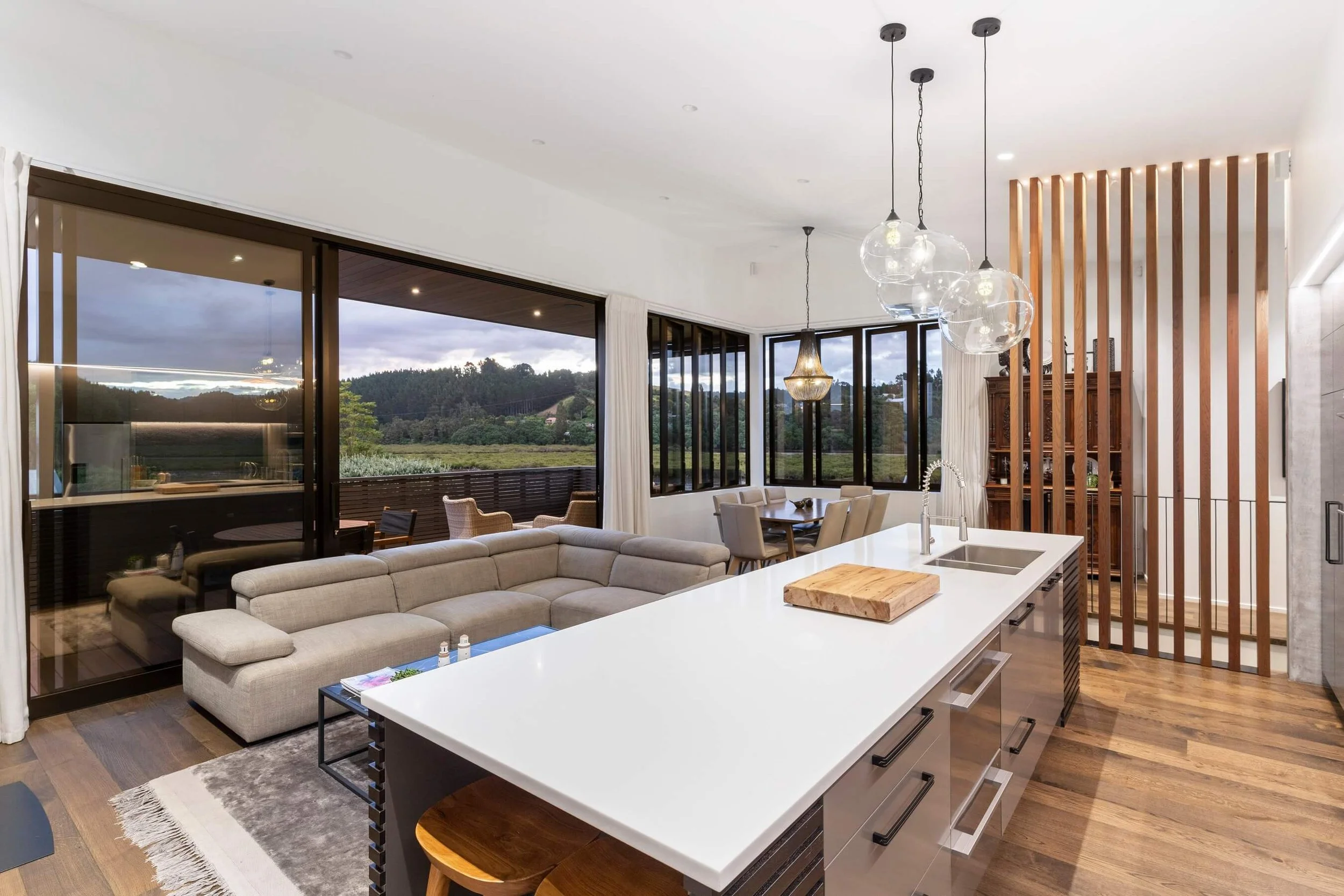 Modern open-concept living room and kitchen with large windows overlooking green hills, featuring a beige sectional sofa, white island with sink, contemporary lighting, and wooden accents.