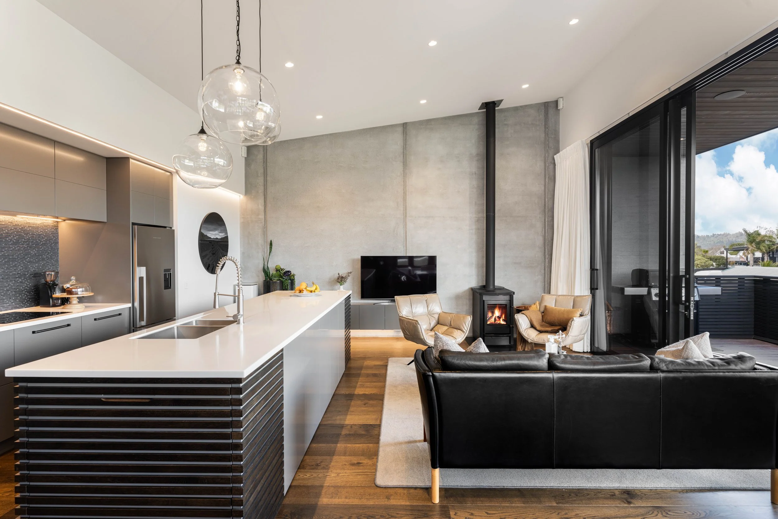 Modern open-concept living space with a kitchen island, leather armchairs near a wood-burning stove, a TV, and large sliding glass doors leading to an outdoor patio, featuring contemporary decor and a concrete accent wall.