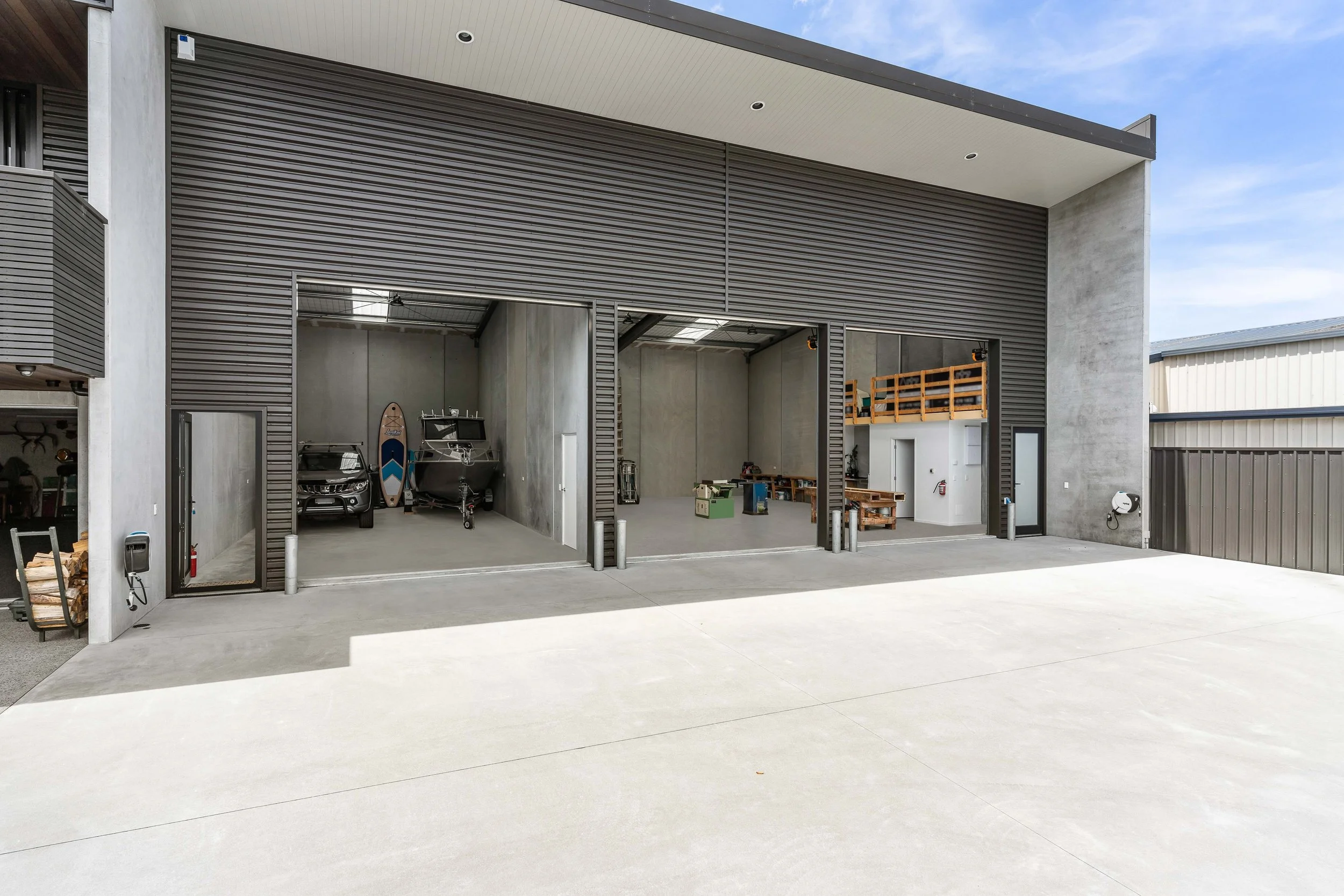 Modern garage with two large open doors, concrete interior, and minimal clutter. Items inside include a boat, a car, and some storage shelves. Exterior features a concrete driveway and a building with a gray and black facade.