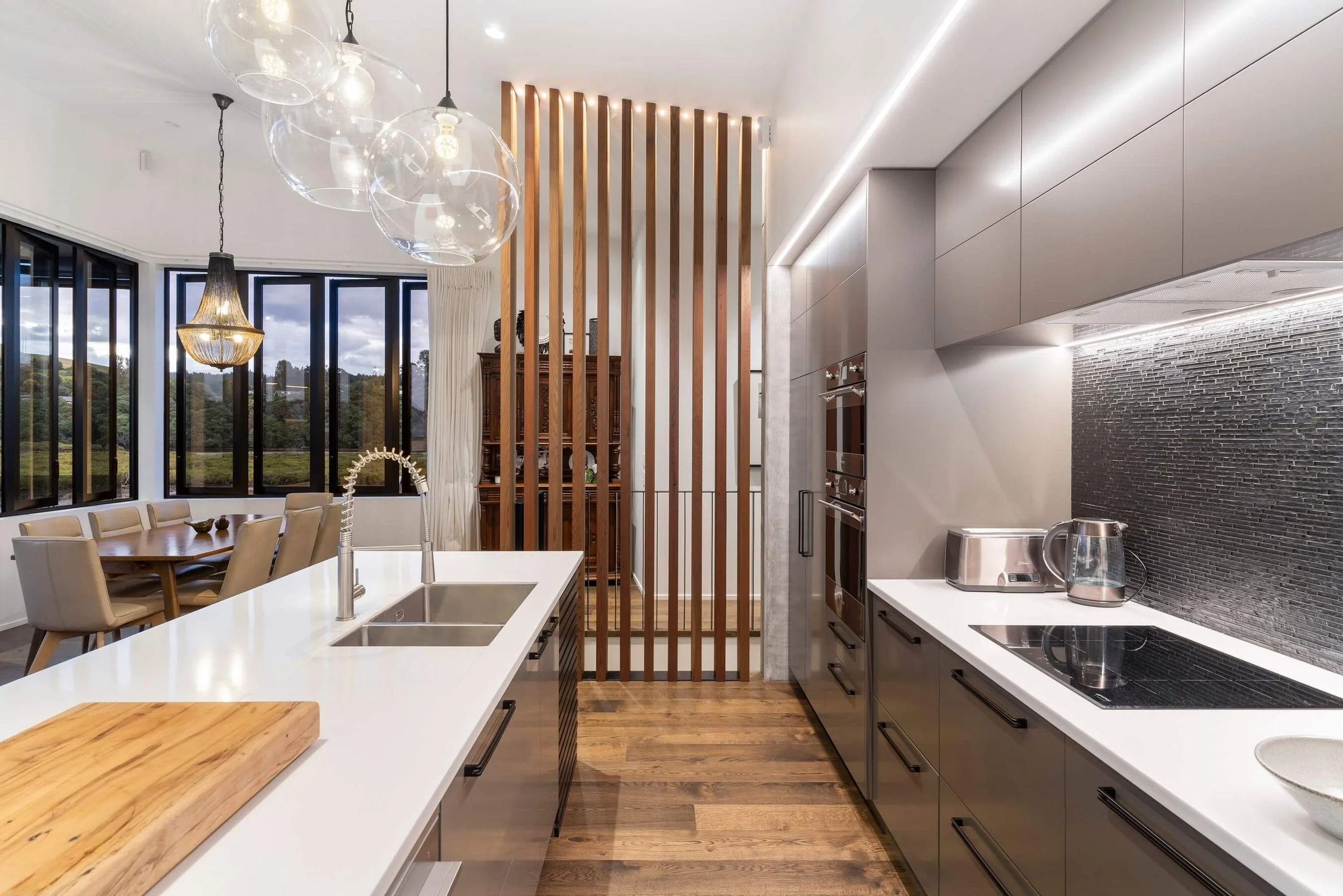 Modern kitchen with white countertops, gray cabinets, black hardware, black tile backsplash, and wooden flooring. Open dining area with large windows, beige chairs, and a wooden dining table. Decorative wooden slats separate the kitchen from the livi