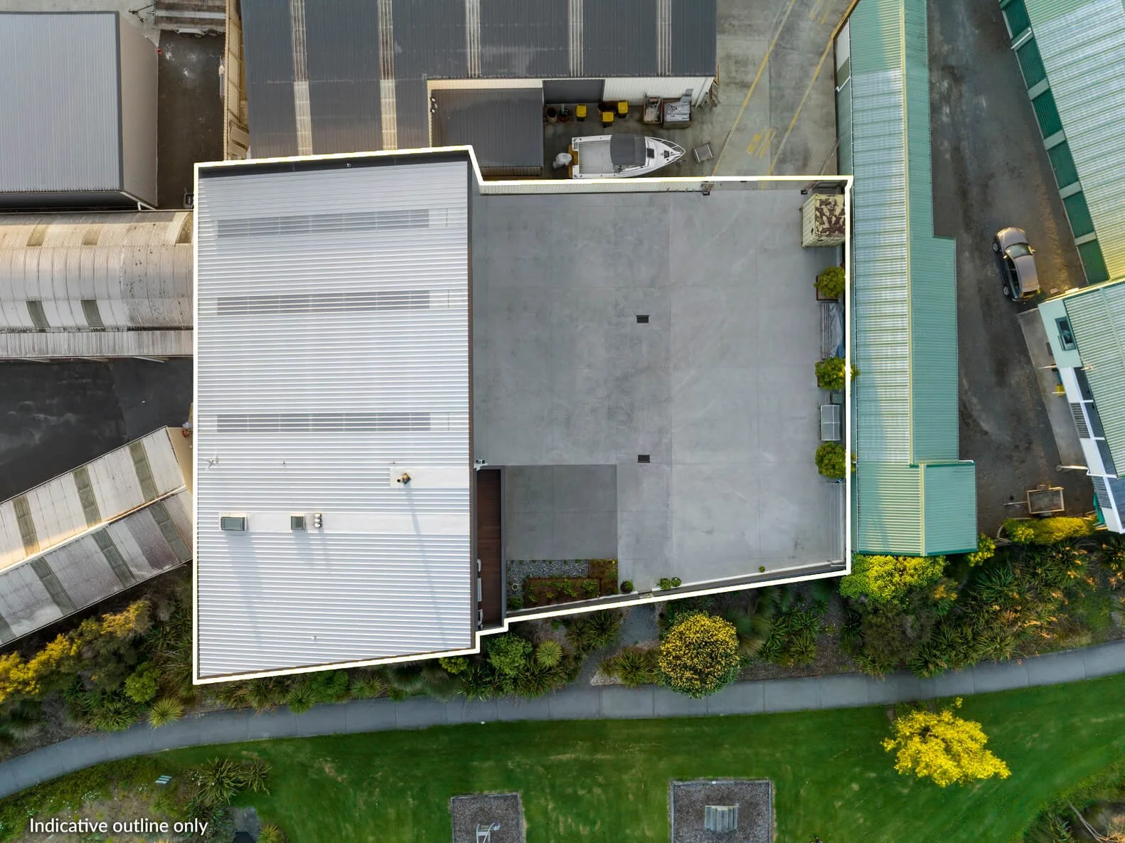 Aerial view of a large flat rooftop with some greenery at the edges, surrounded by neighboring buildings, parking lots, and landscaped areas with trees and grass.