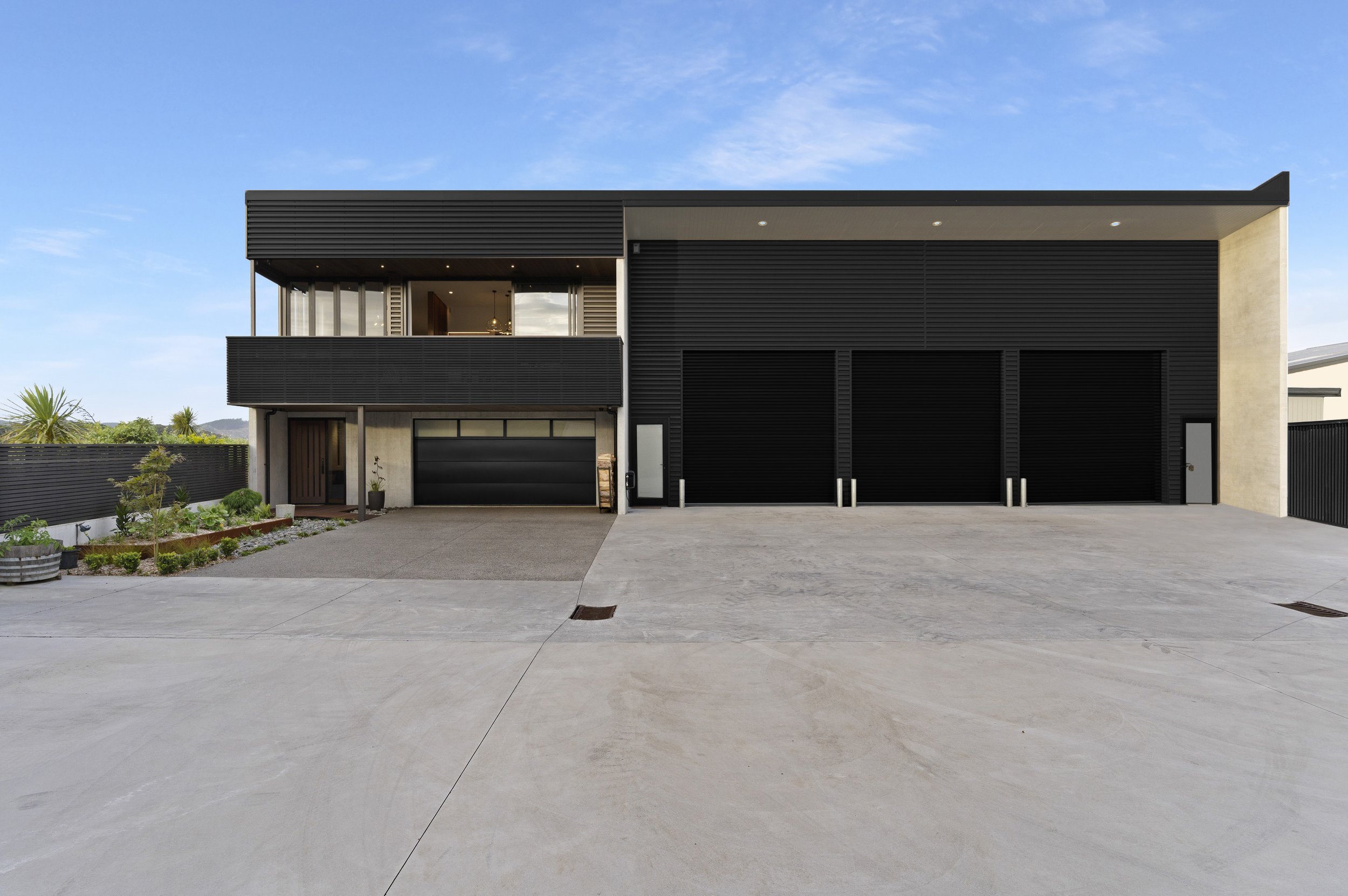 Front view of a modern two-story house with a black and gray exterior, garage doors, small front yard, and clear blue sky.