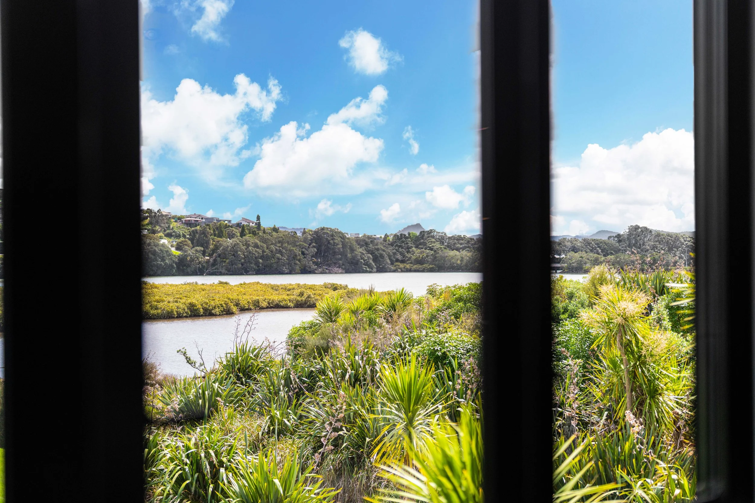 View of a blue sky with clouds through black window bars, overlooking the Moana Anu estuary, trees, and houses on a hill.