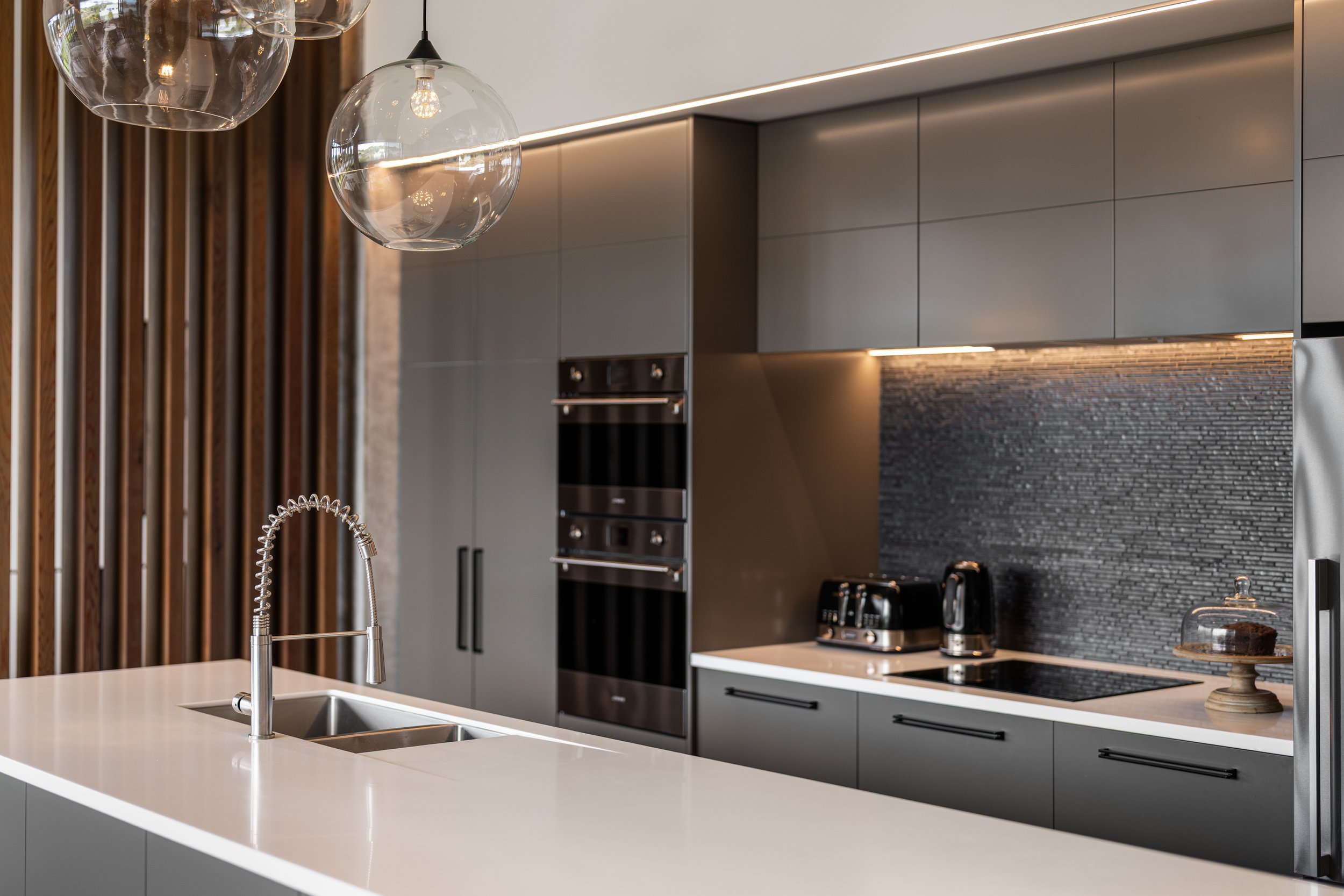 Modern kitchen with gray cabinets, black backsplash, and a white countertop island with a sink and a high-arched faucet. Glass pendant lights hang above the island.