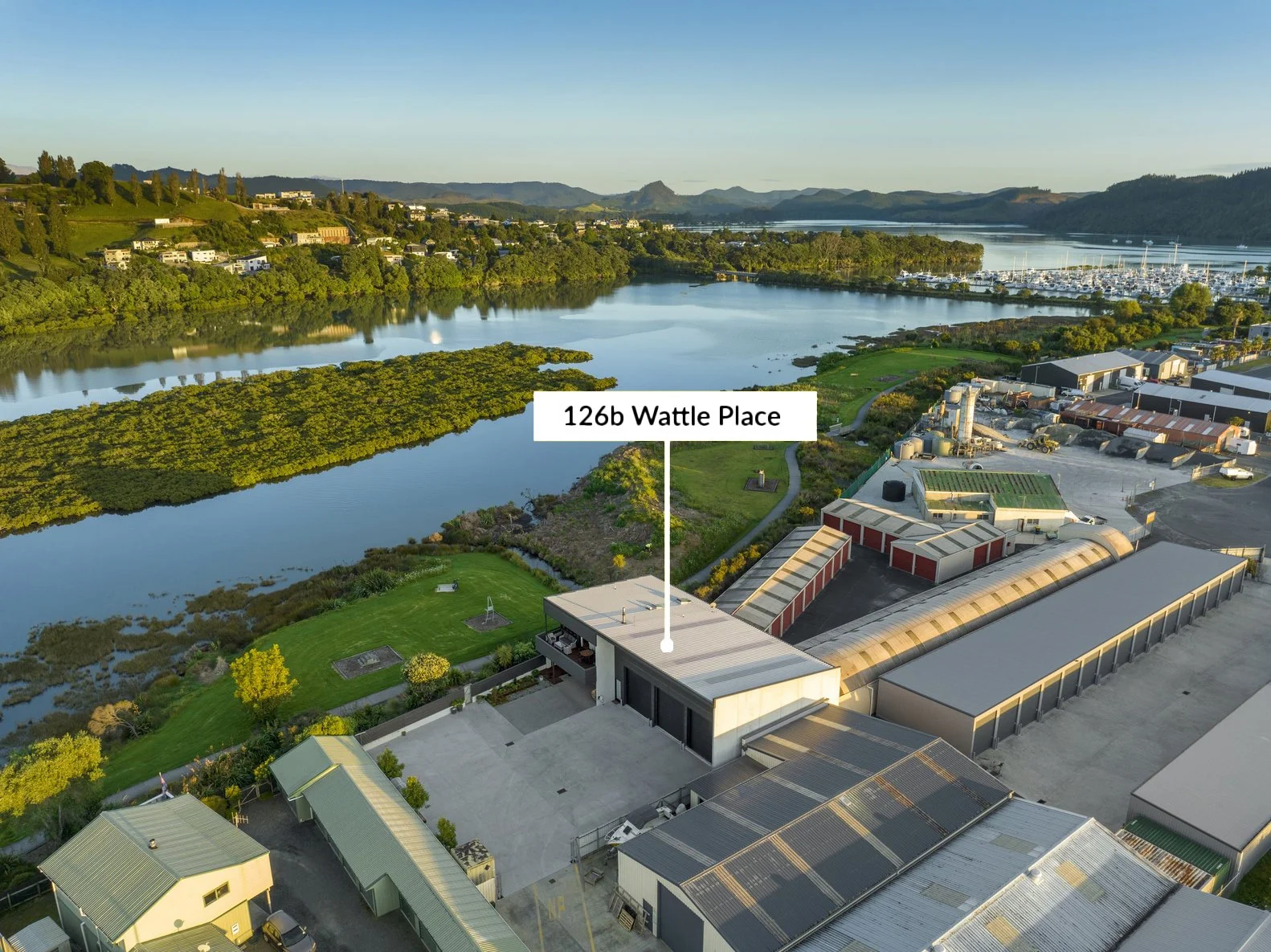 Aerial view of a riverside area with a marina, residential buildings, and industrial warehouses labeled with a flag and name, water, trees, and hilly landscape in the background.