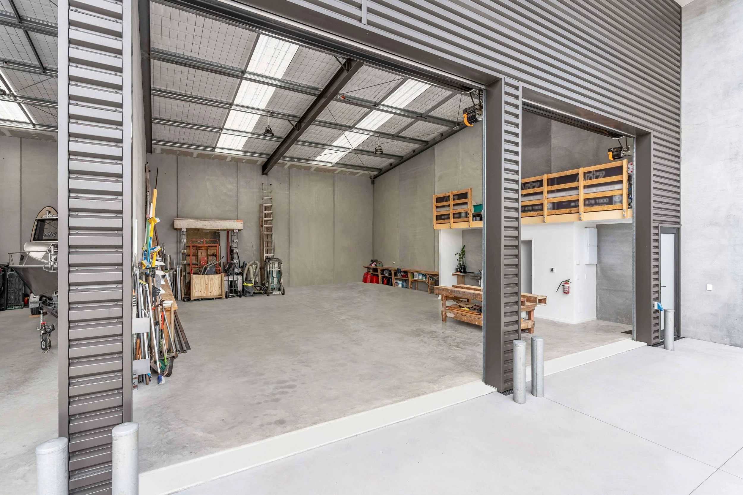 Empty industrial garage with metal sliding doors open, showing a concrete floor, storage shelves, ladders, and tools inside.