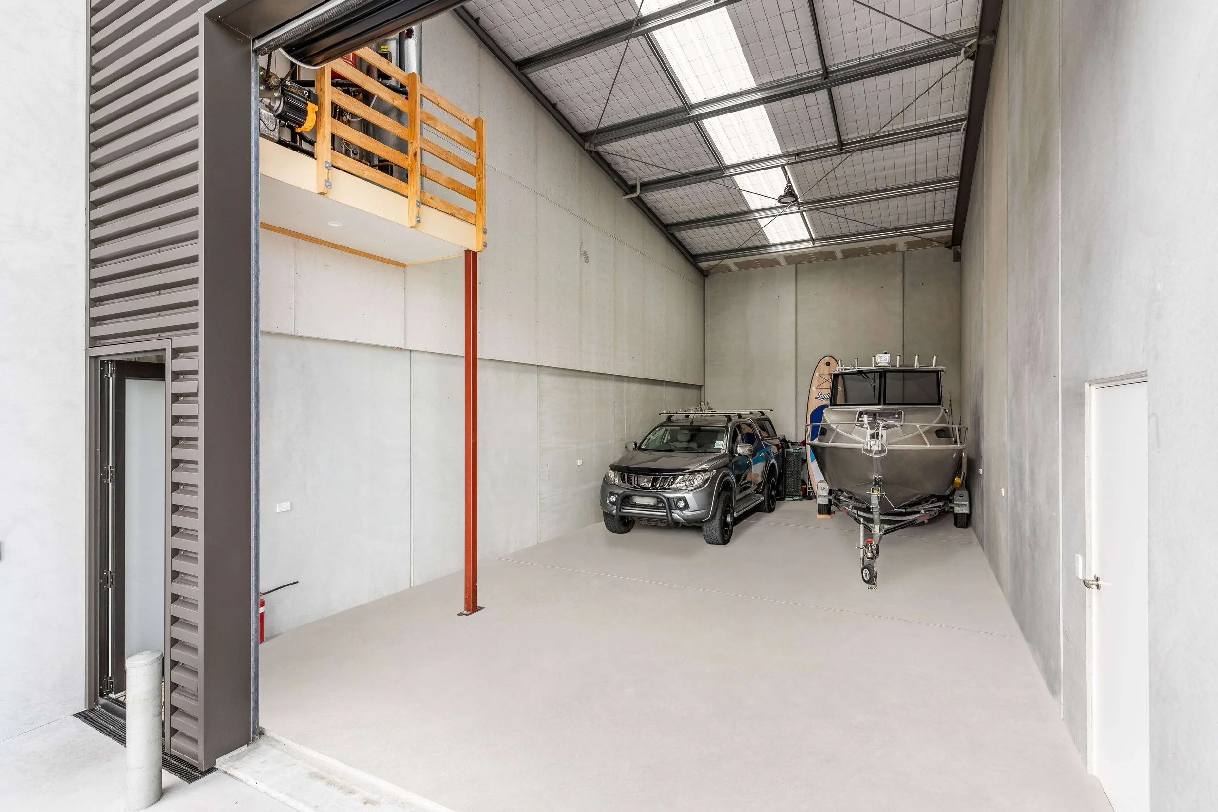Large indoor space with a high ceiling and concrete walls. Inside are a gray SUV, a boat on a trailer, and a small wooden loft with a railing. A vertical red pole supports the loft, and there is a black ventilation or air conditioning unit on the lef