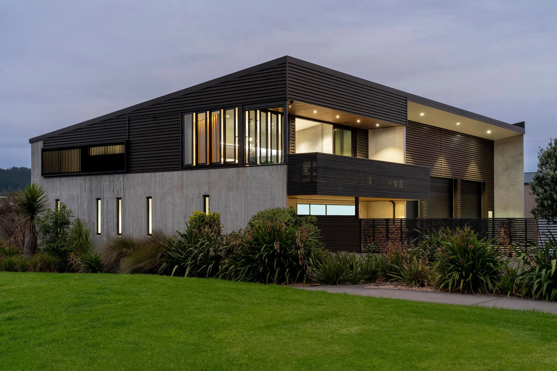 Modern two-story house with a flat roof, black horizontal slats, and large windows, surrounded by a well-maintained lawn and plants, during dusk with interior lights on.