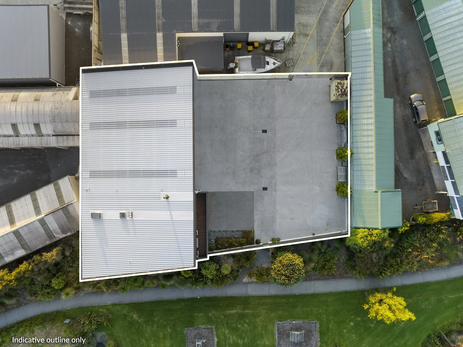 From above, an aerial view shows a building with a white metal roof, possibly commercial or industrial, with rooftop vents and HVAC units. Surrounding the building are neighboring structures, parking areas, and greenery, including a landscaped sectio