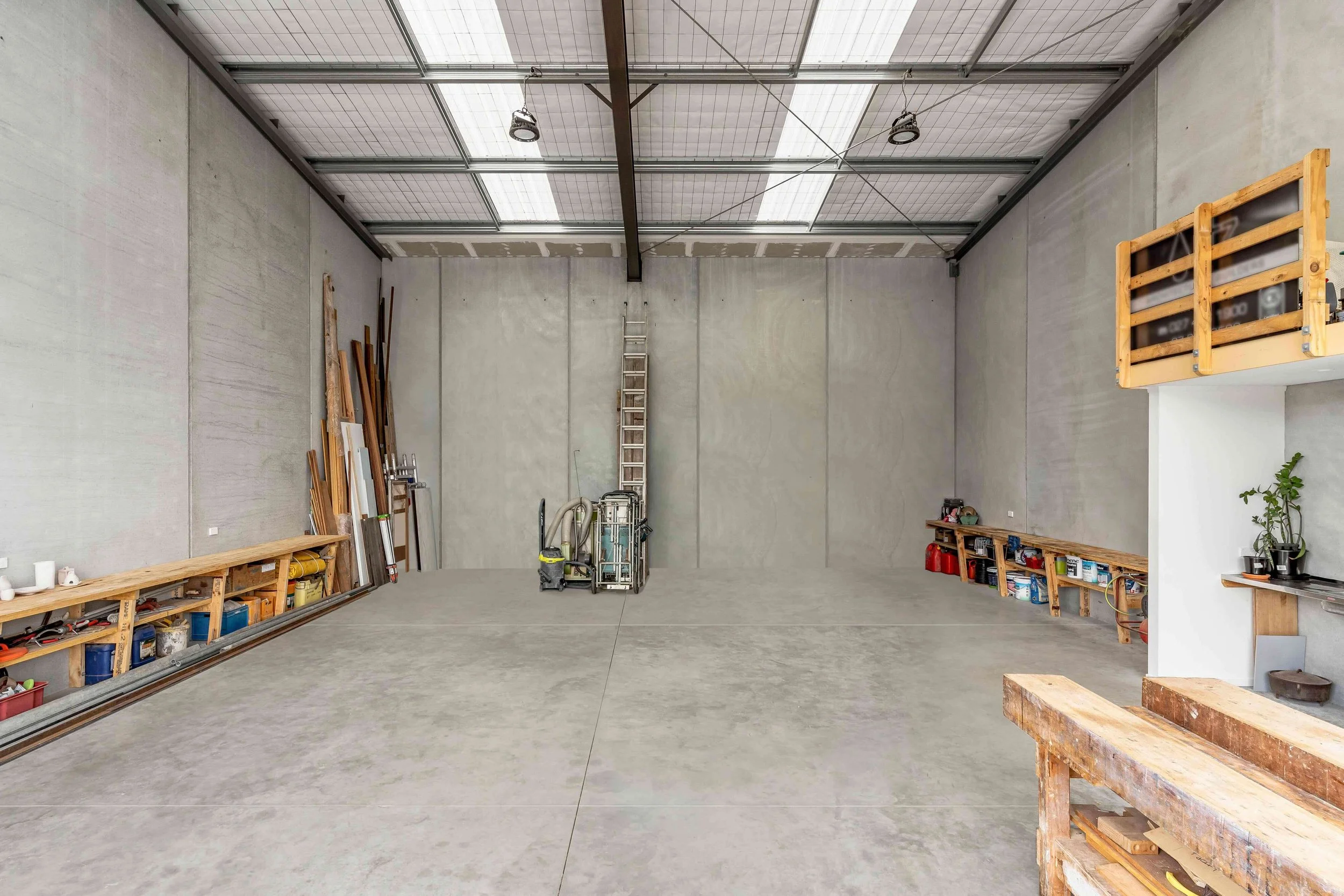 Empty garage or workshop with tools, wooden shelves, and a ladder against the wall, concrete floor and ceiling with lights.