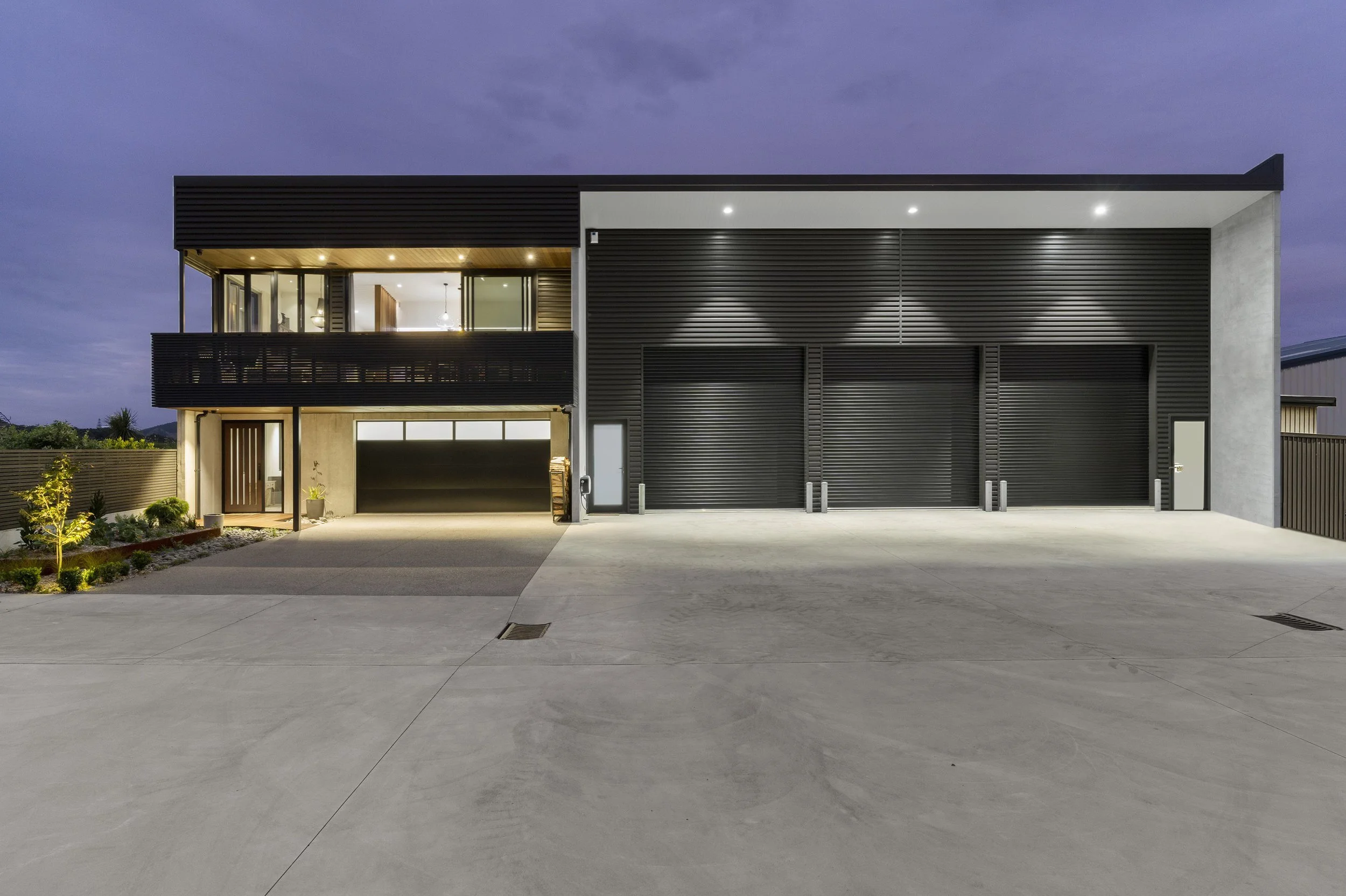 Modern two-story house with black exterior, large garage, and illuminated interior at dusk. Concrete driveway and small landscaping in front. Clear evening sky.